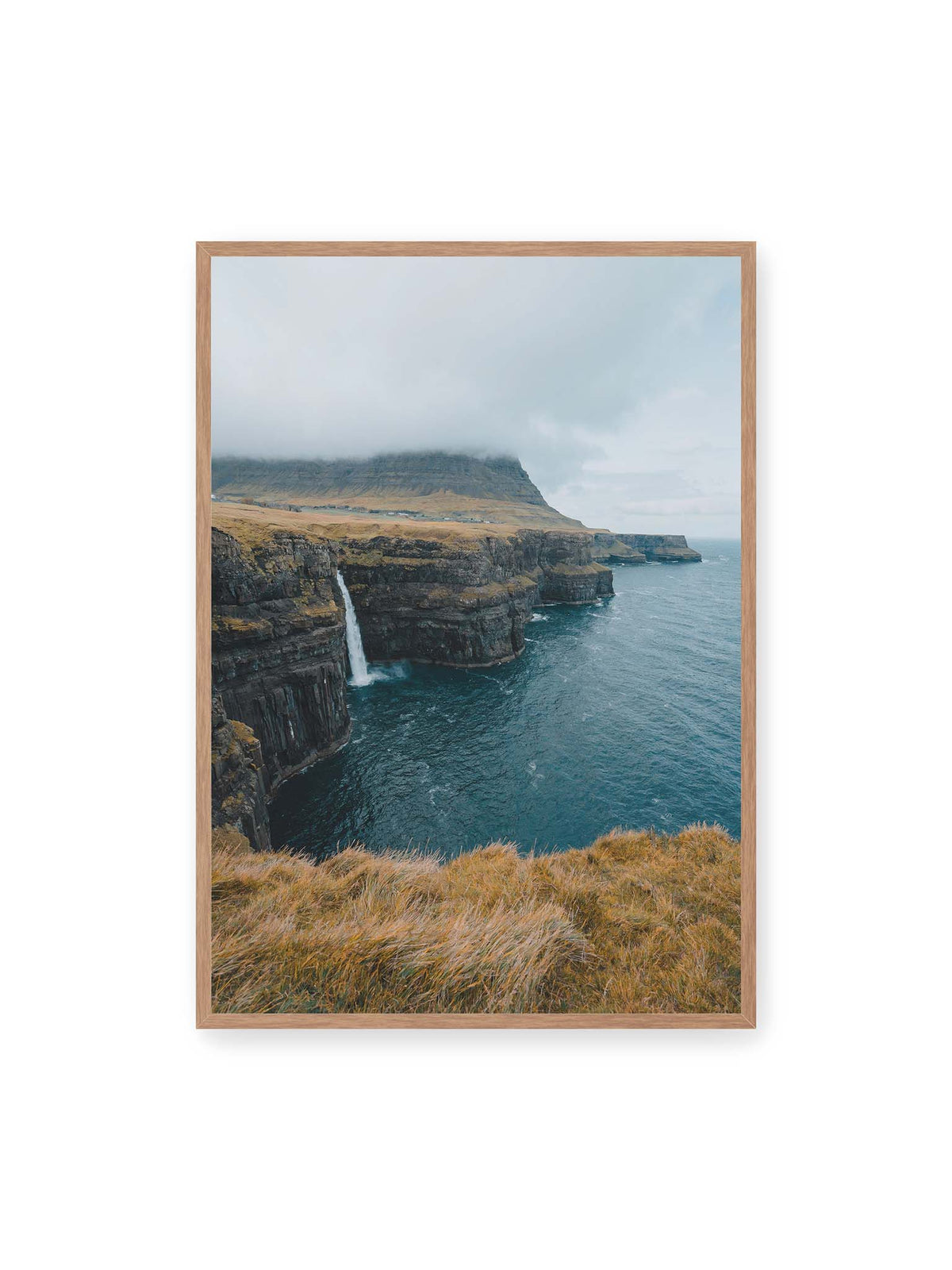70x100 cm, Eichenrahmen – Dunkle Küstenklippen mit Wasserfall, grasigem Hochland und tief hängenden Wolken über blauem Meer.