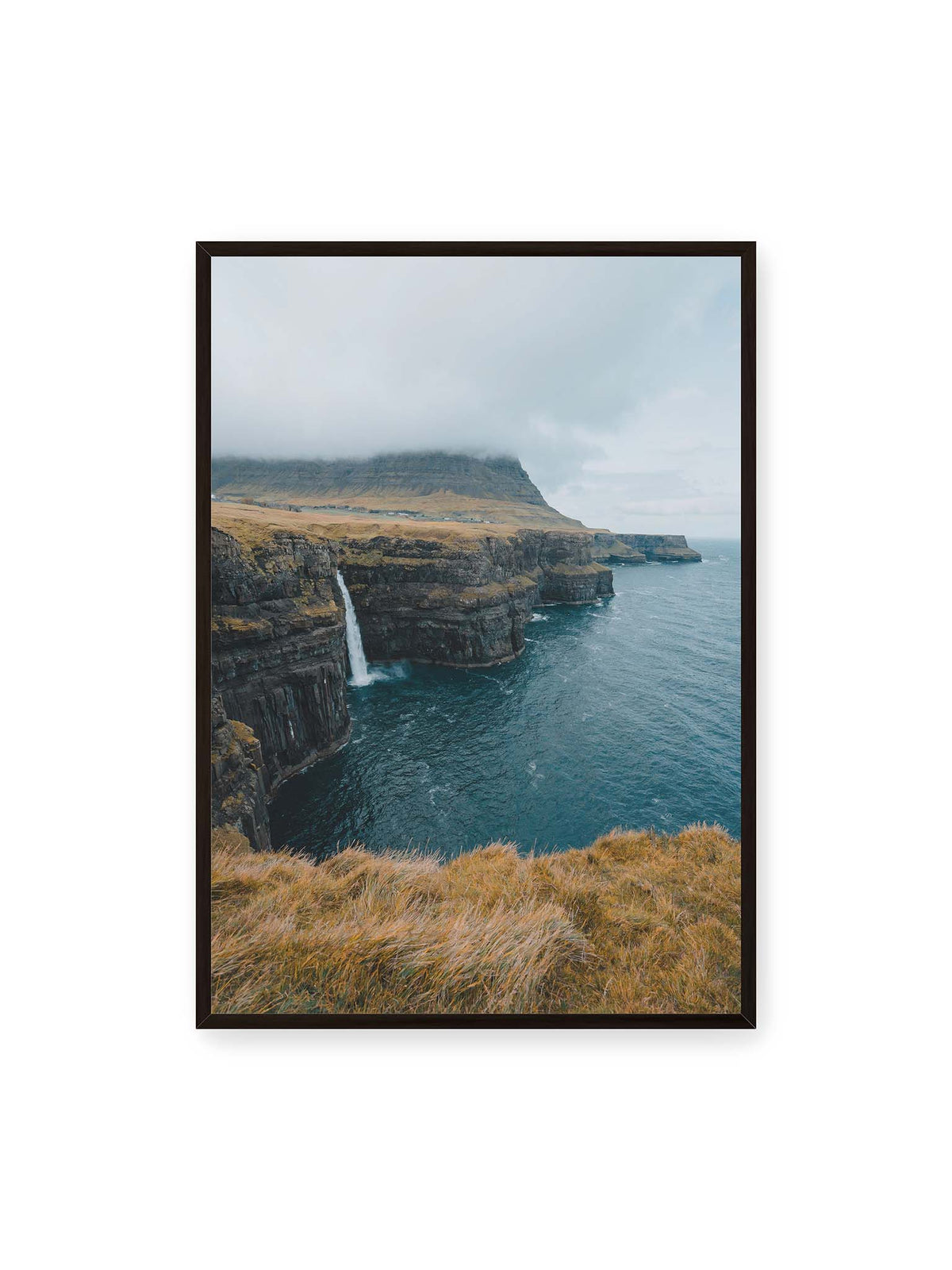 70x100 cm, schwarzer Rahmen – Dunkle Küstenklippen mit Wasserfall, grasigem Hochland und tief hängenden Wolken über blauem Meer.