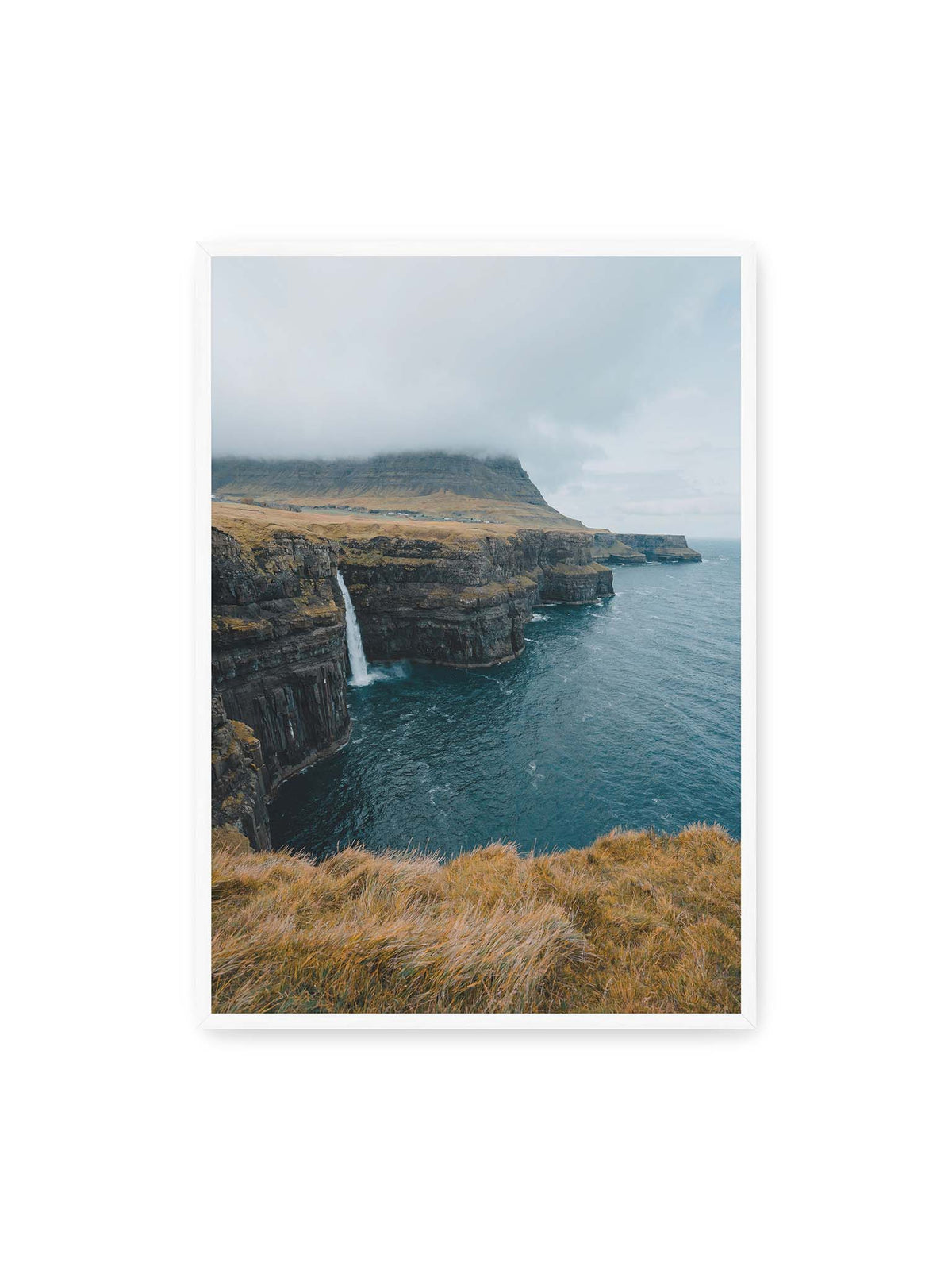 70x100 cm, weißer Rahmen – Dunkle Küstenklippen mit Wasserfall, grasigem Hochland und tief hängenden Wolken über blauem Meer.