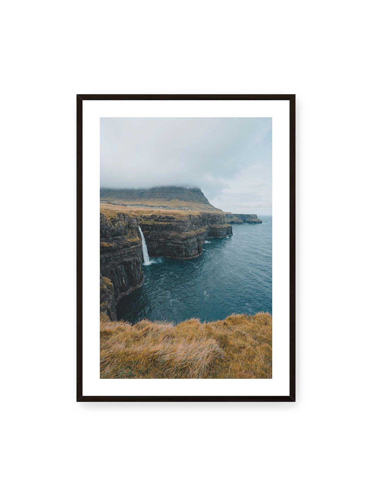 70x100 cm, weißer Galerie-Rand, schwarzer Rahmen – Dunkle Küstenklippen mit Wasserfall, grasigem Hochland und tief hängenden Wolken über blauem Meer.
