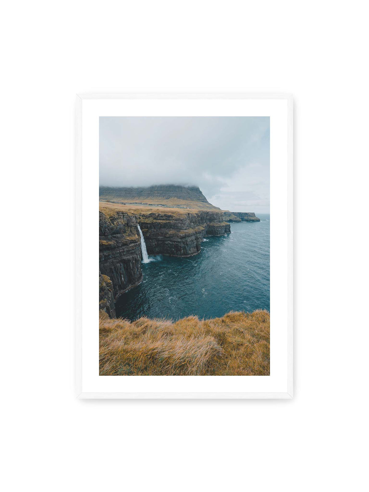 70x100 cm, weißer Galerie-Rand, weißer Rahmen – Dunkle Küstenklippen mit Wasserfall, grasigem Hochland und tief hängenden Wolken über blauem Meer.
