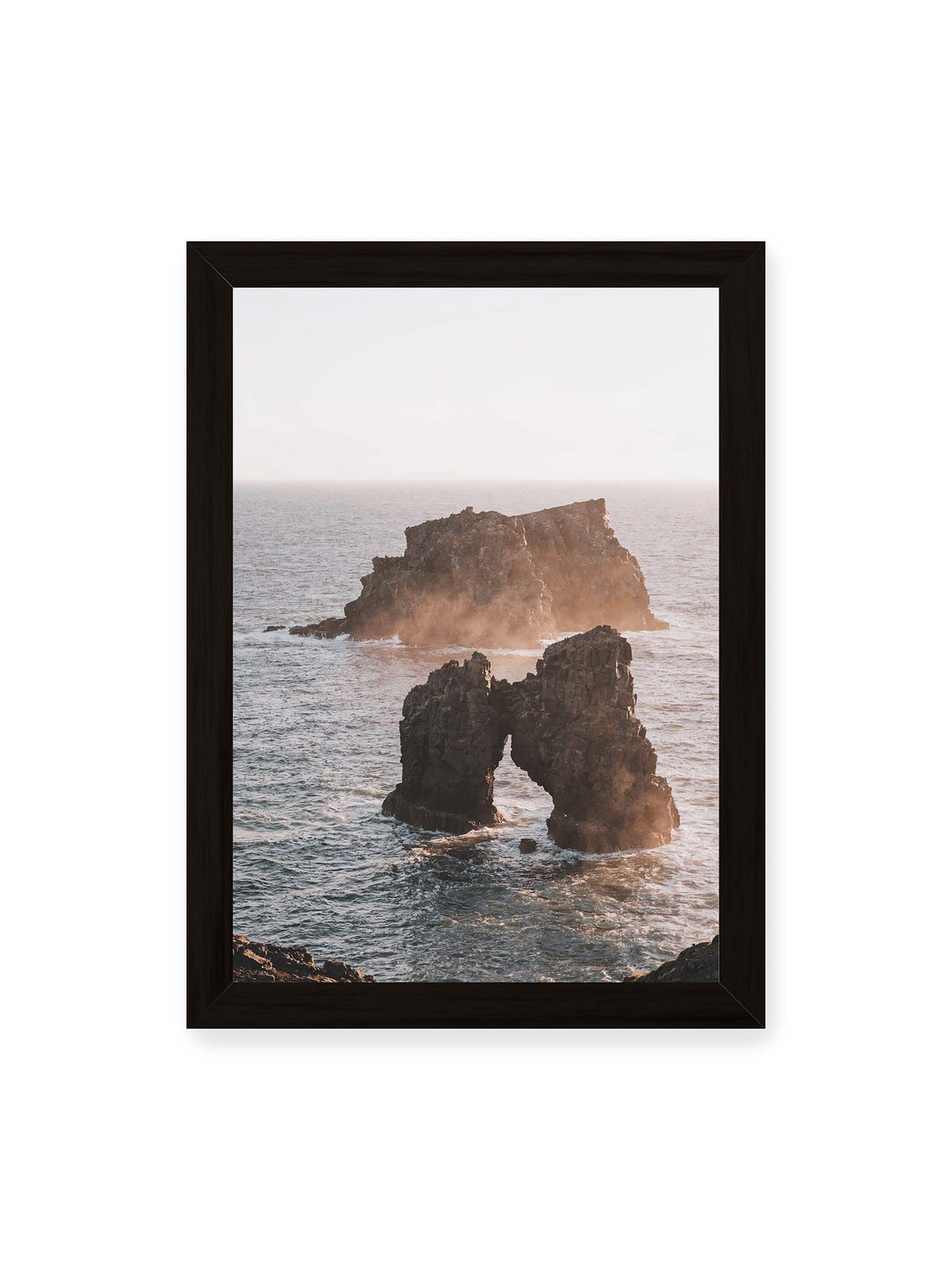 21x30 cm, schwarzer Rahmen – Dunkler Felsbogen und massive Felsformation im bewegten Meer, umgeben von heller Gischt und warmem Licht.