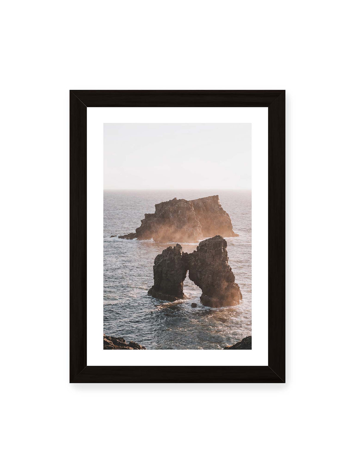 21x30 cm, weißer Galerie-Rand, schwarzer Rahmen – Dunkler Felsbogen und massive Felsformation im bewegten Meer, umgeben von heller Gischt und warmem Licht.