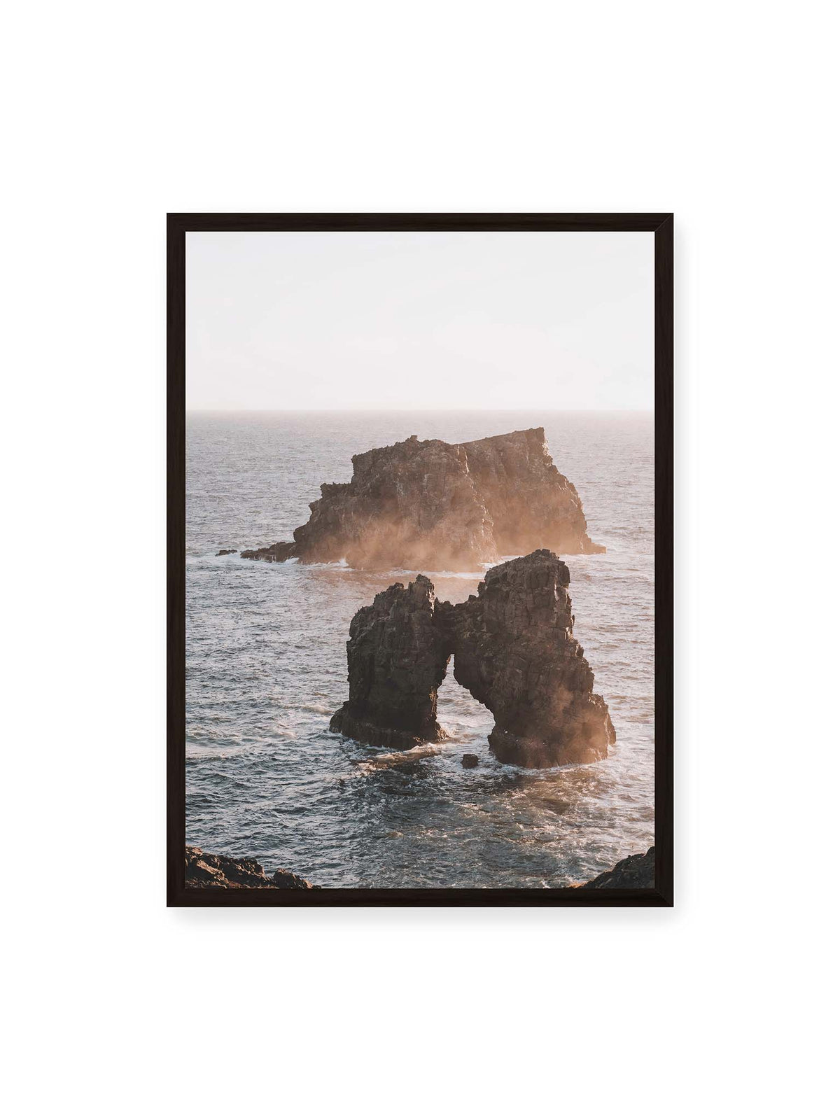 50x70 cm, schwarzer Rahmen – Dunkler Felsbogen und massive Felsformation im bewegten Meer, umgeben von heller Gischt und warmem Licht.