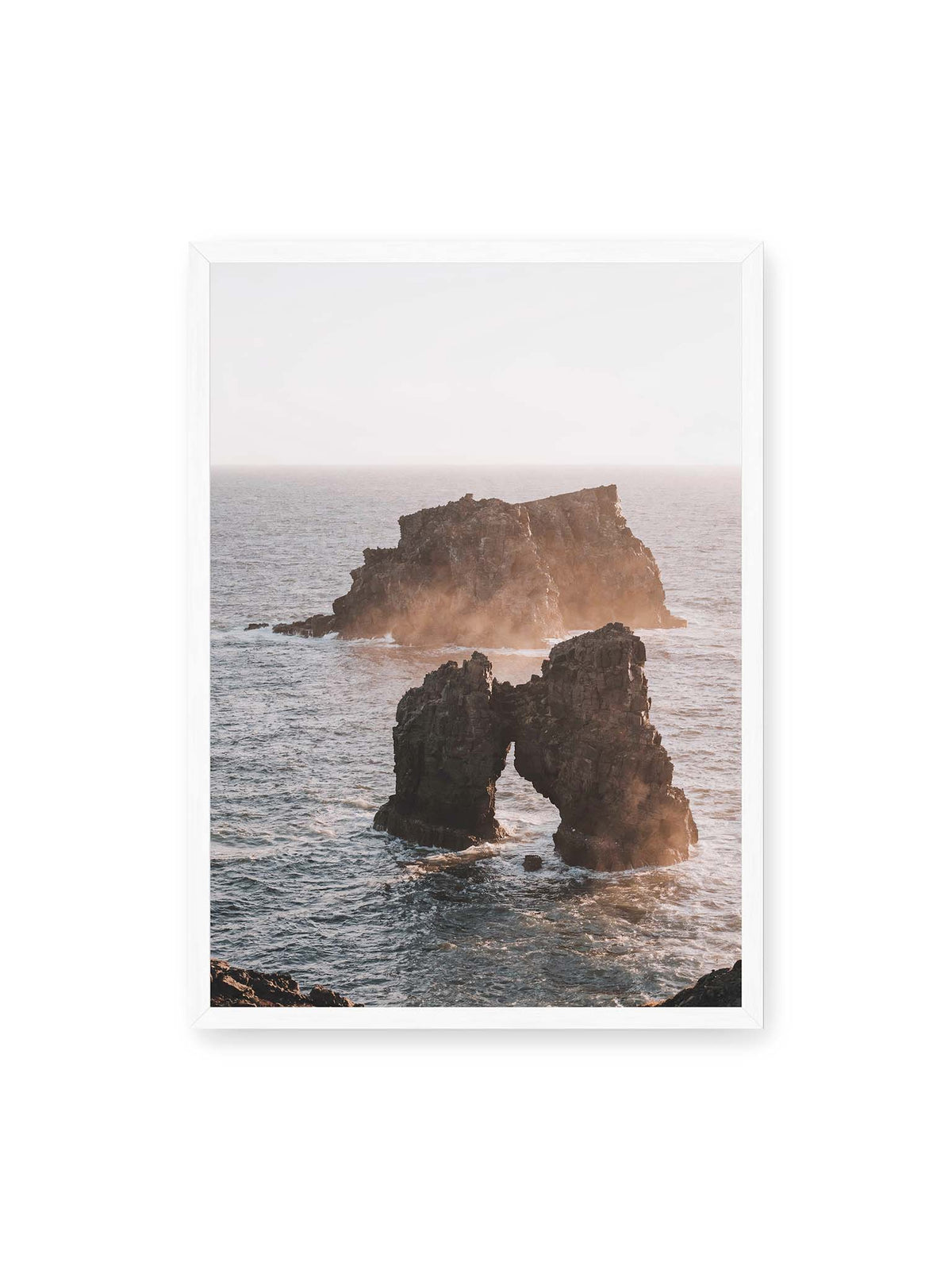 50x70 cm, weißer Rahmen – Dunkler Felsbogen und massive Felsformation im bewegten Meer, umgeben von heller Gischt und warmem Licht.