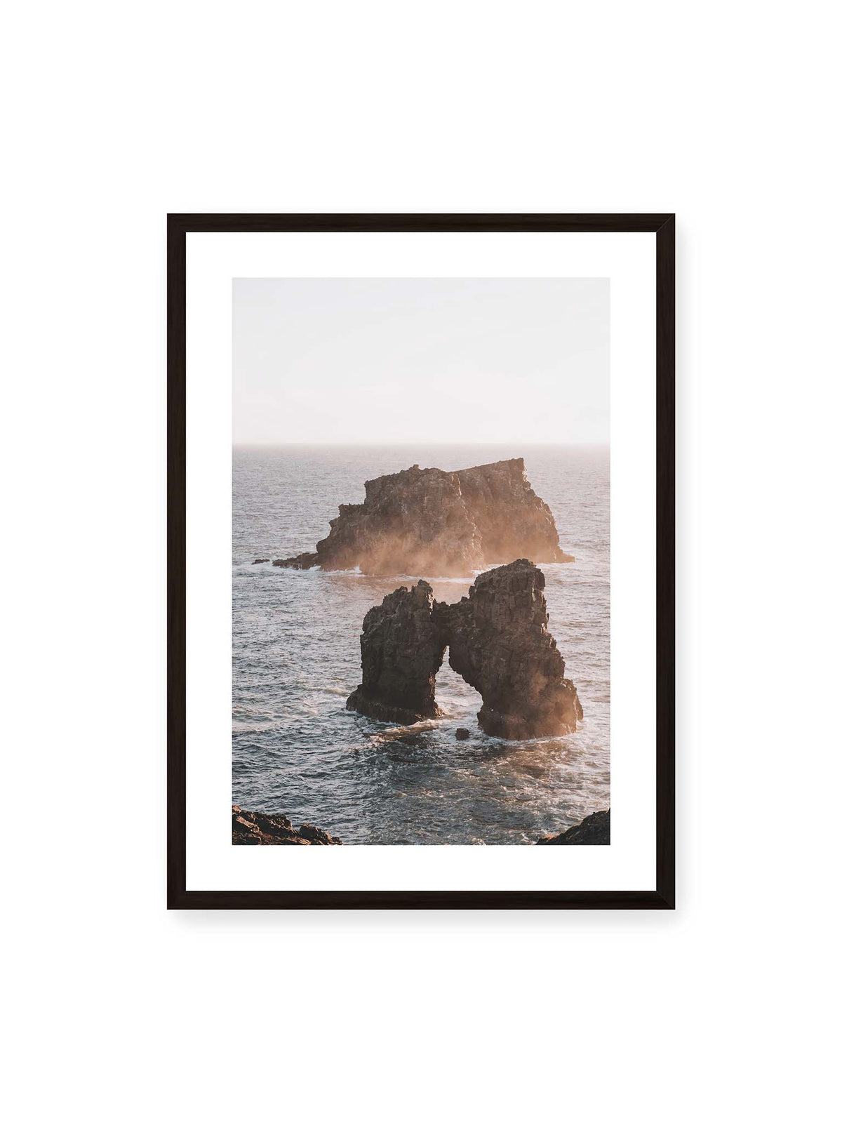 50x70 cm, weißer Galerie-Rand, schwarzer Rahmen – Dunkler Felsbogen und massive Felsformation im bewegten Meer, umgeben von heller Gischt und warmem Licht.