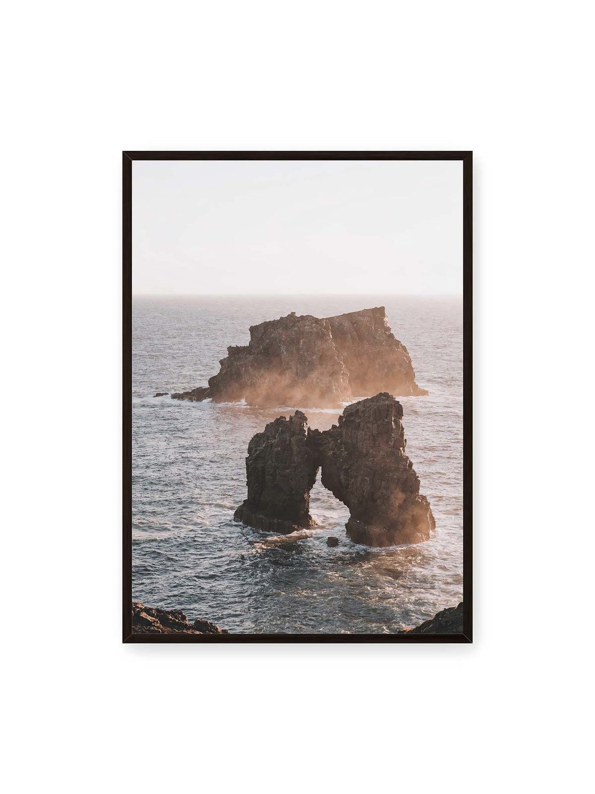 70x100 cm, schwarzer Rahmen – Dunkler Felsbogen und massive Felsformation im bewegten Meer, umgeben von heller Gischt und warmem Licht.