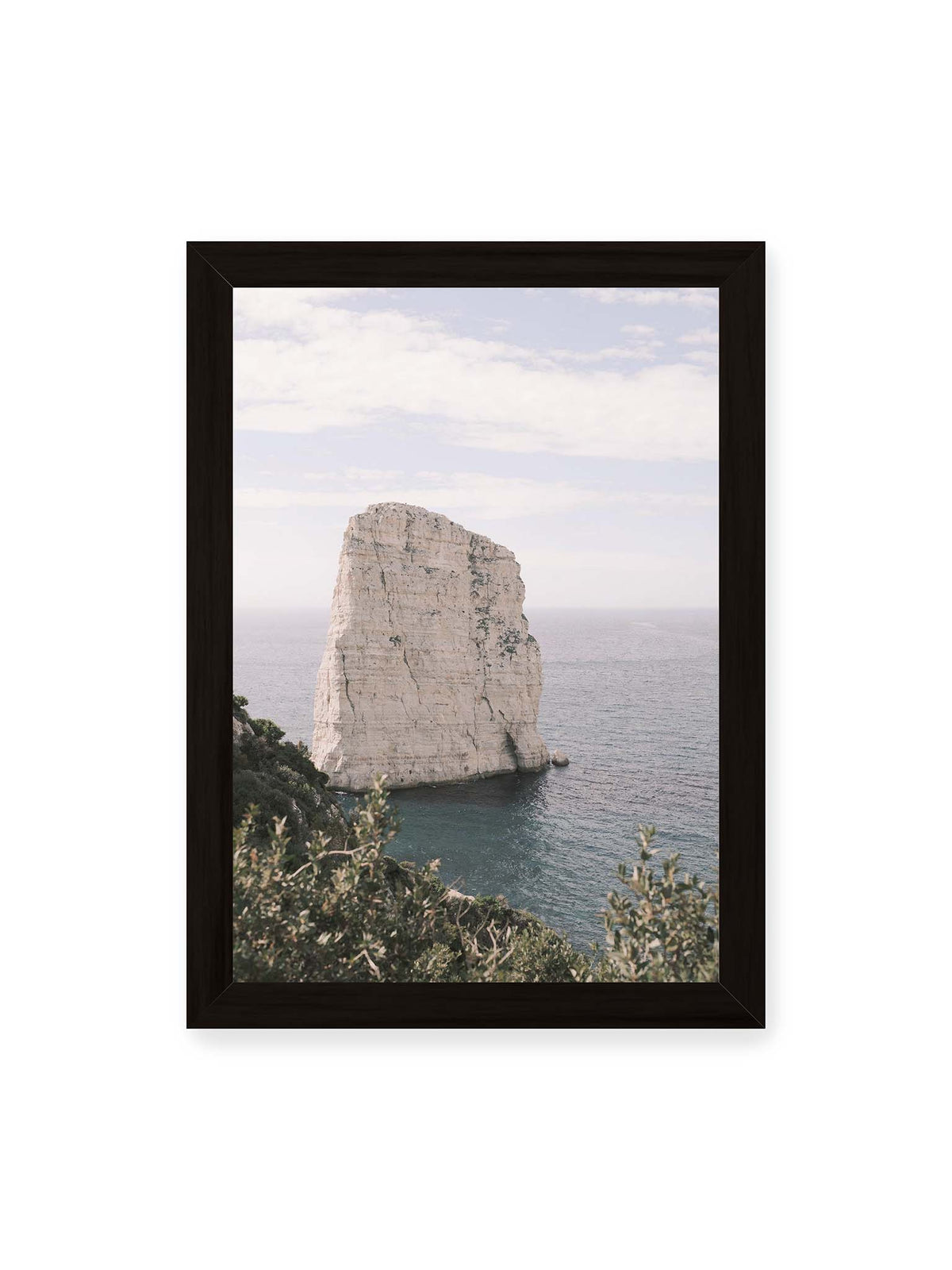 21x30 cm, schwarzer Rahmen – Heller, steil aufragender Felsen im Meer vor blassem Himmel, eingerahmt von grüner Vegetation.