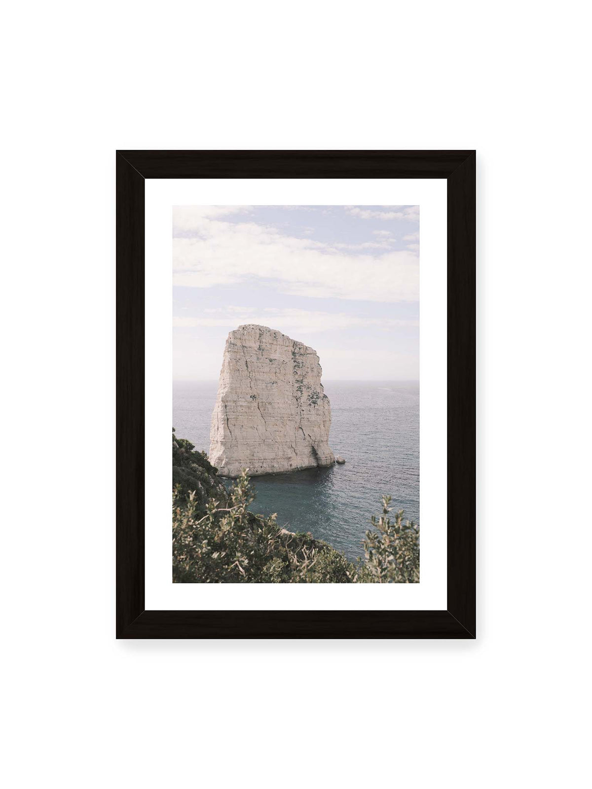 21x30 cm, weißer Galerie-Rand, schwarzer Rahmen – Heller, steil aufragender Felsen im Meer vor blassem Himmel, eingerahmt von grüner Vegetation.