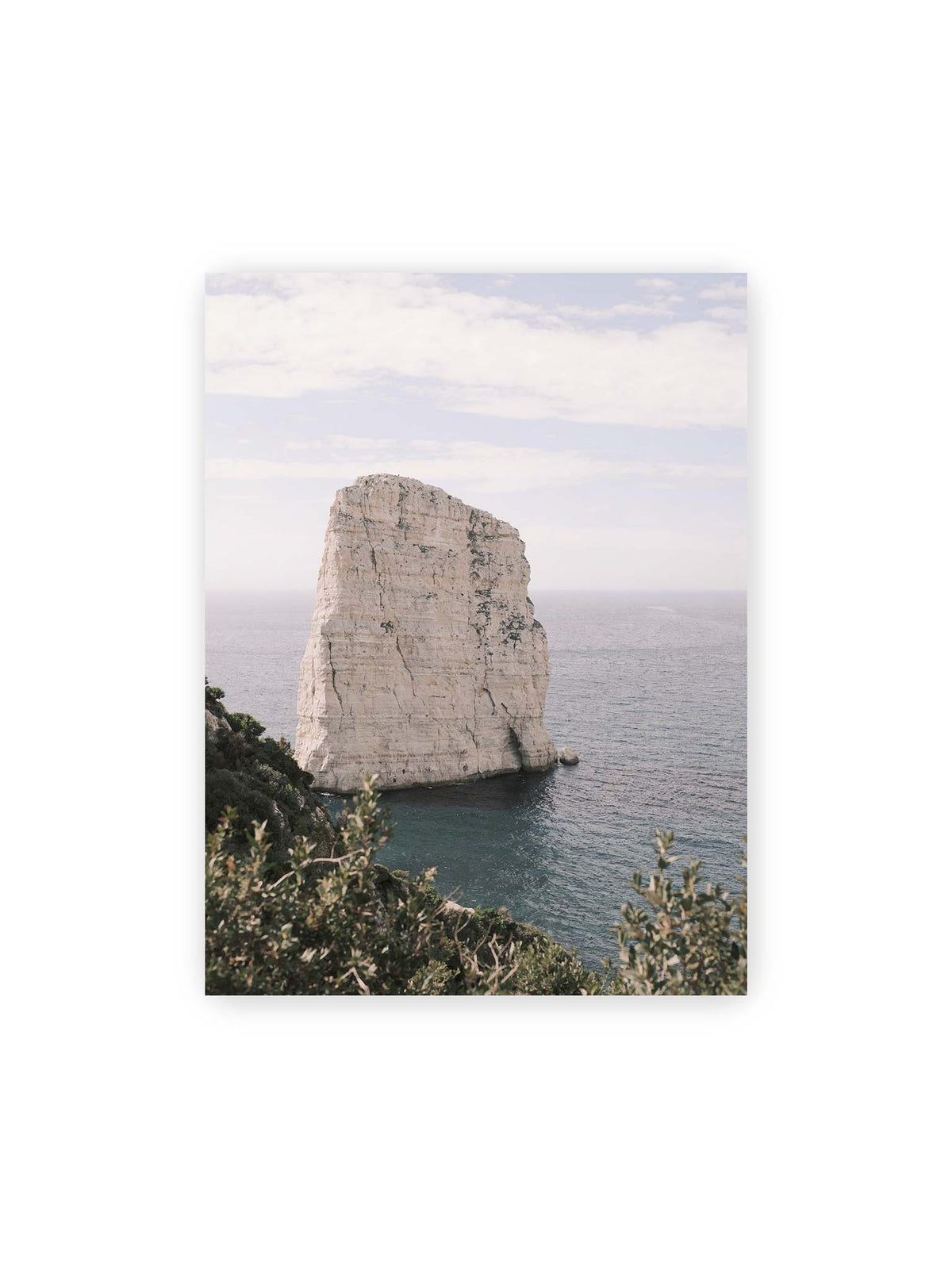 30x40 cm – Heller, steil aufragender Felsen im Meer vor blassem Himmel, eingerahmt von grüner Vegetation.