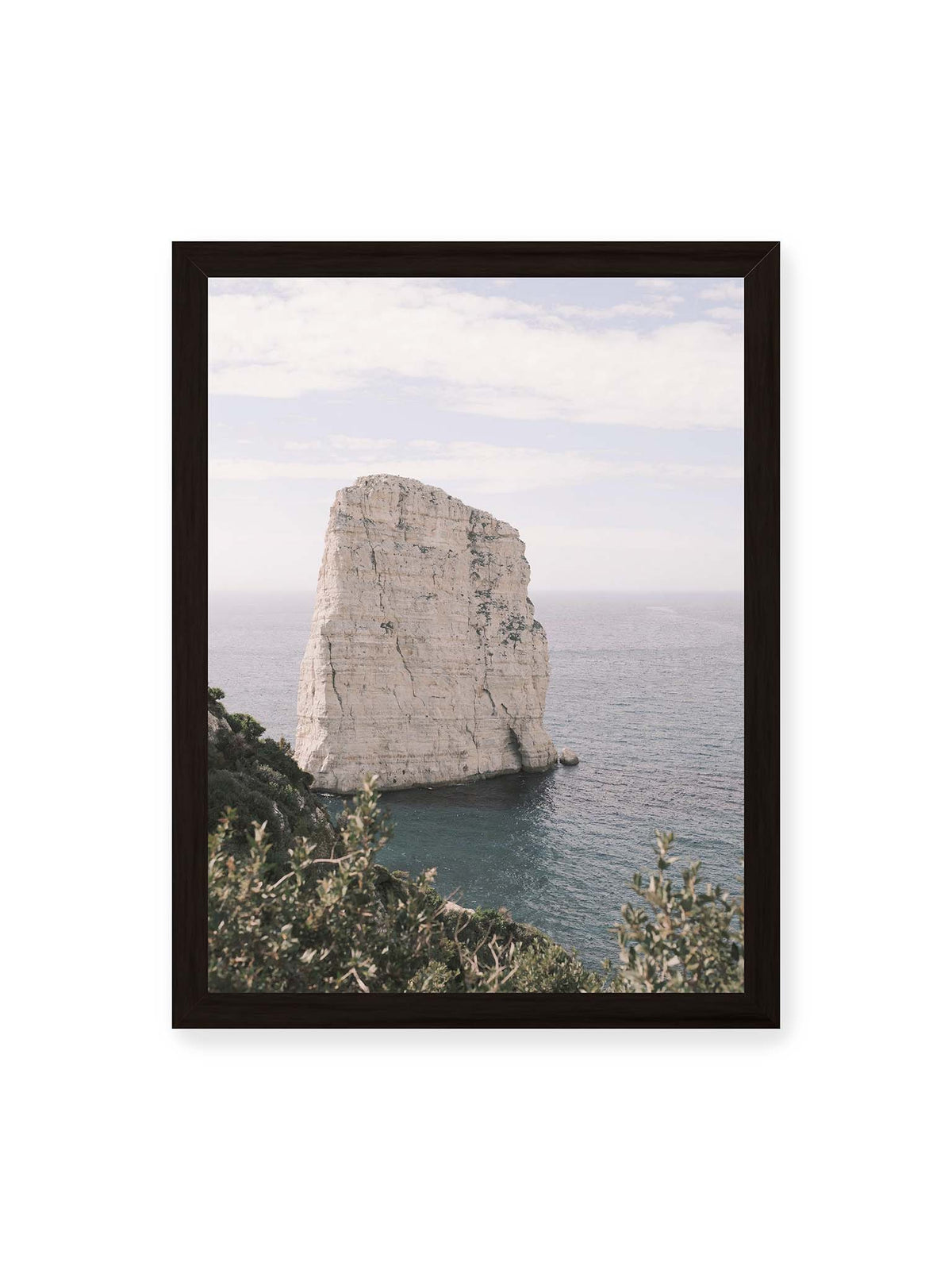 30x40 cm, schwarzer Rahmen – Heller, steil aufragender Felsen im Meer vor blassem Himmel, eingerahmt von grüner Vegetation.