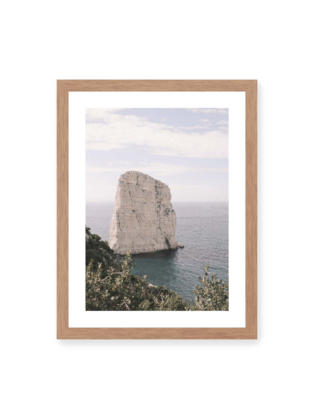 30x40 cm, weißer Galerie-Rand, Eichenrahmen – Heller, steil aufragender Felsen im Meer vor blassem Himmel, eingerahmt von grüner Vegetation.