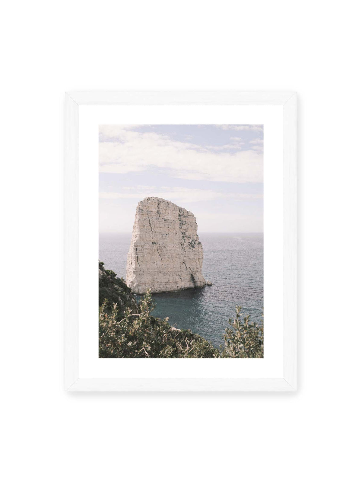30x40 cm, weißer Galerie-Rand, weißer Rahmen – Heller, steil aufragender Felsen im Meer vor blassem Himmel, eingerahmt von grüner Vegetation.