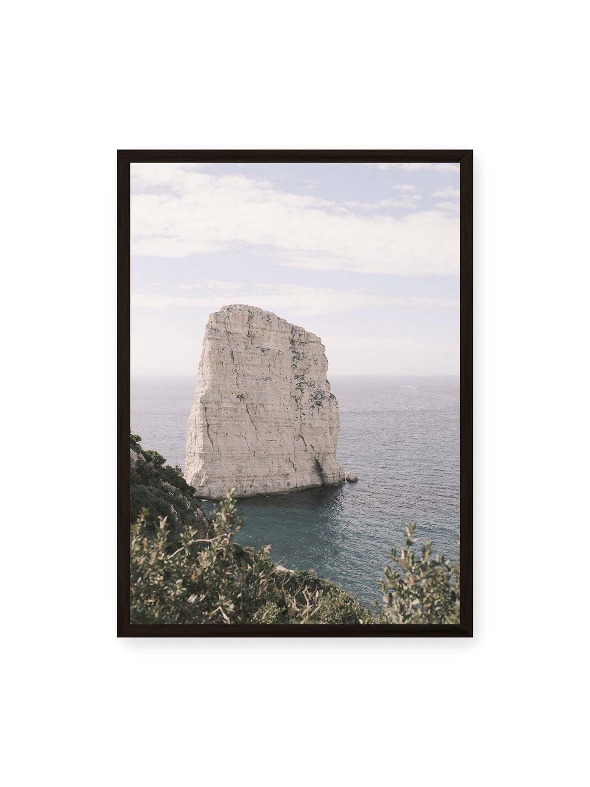 50x70 cm, schwarzer Rahmen – Heller, steil aufragender Felsen im Meer vor blassem Himmel, eingerahmt von grüner Vegetation.