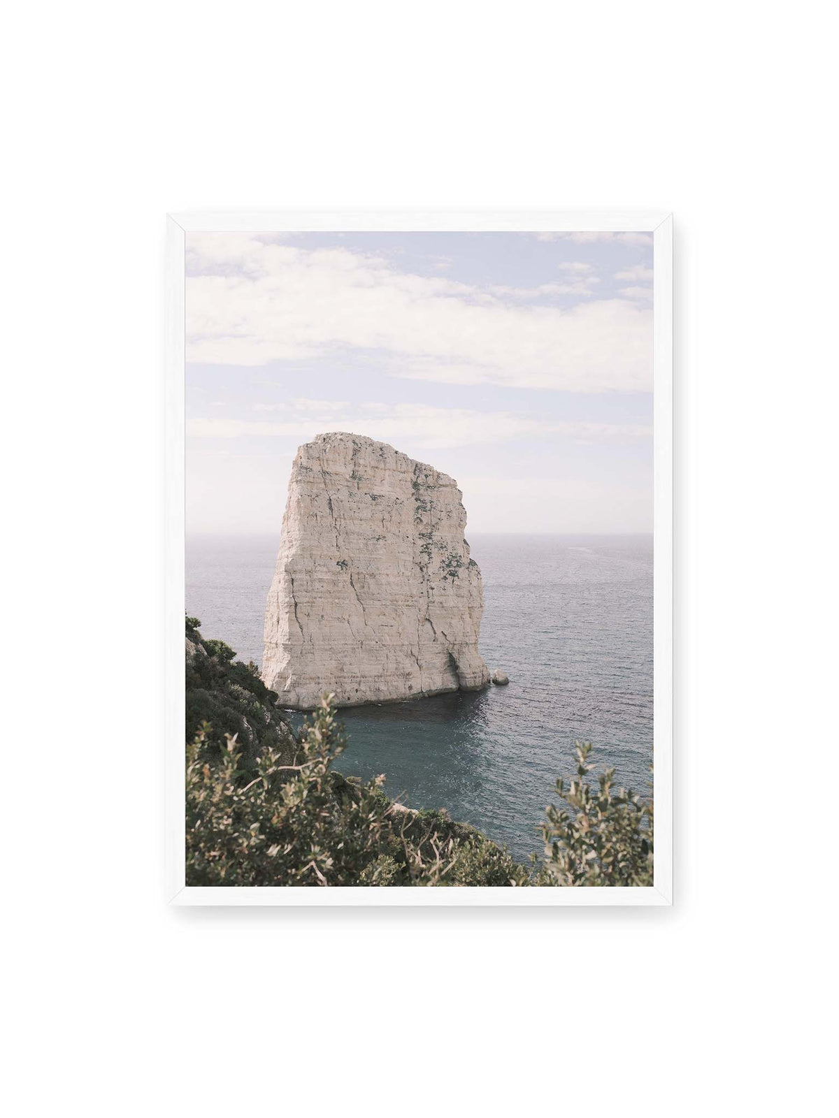 50x70 cm, weißer Rahmen – Heller, steil aufragender Felsen im Meer vor blassem Himmel, eingerahmt von grüner Vegetation.