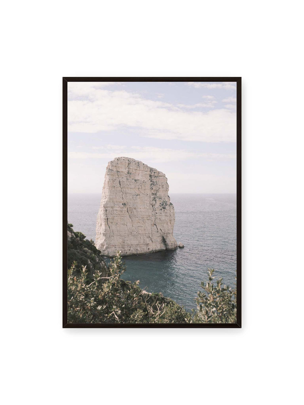 70x100 cm, schwarzer Rahmen – Heller, steil aufragender Felsen im Meer vor blassem Himmel, eingerahmt von grüner Vegetation.