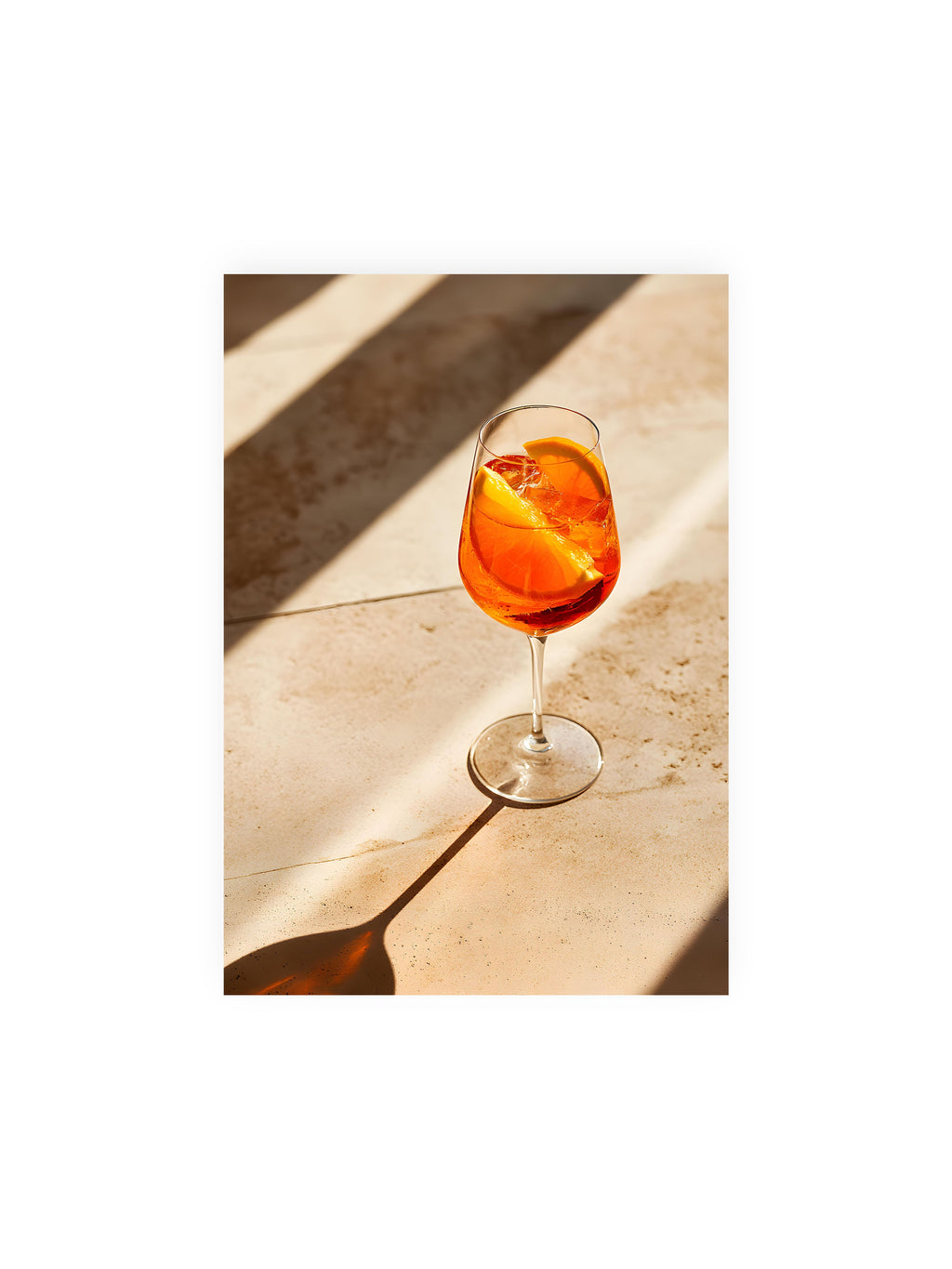 21x30 cm ohne Galerie-Rand ohne Rahmen – Aperitifglas mit orangefarbenem Drink und Orangenscheiben, Sonnenlicht und langer Schatten auf hellem Stein.