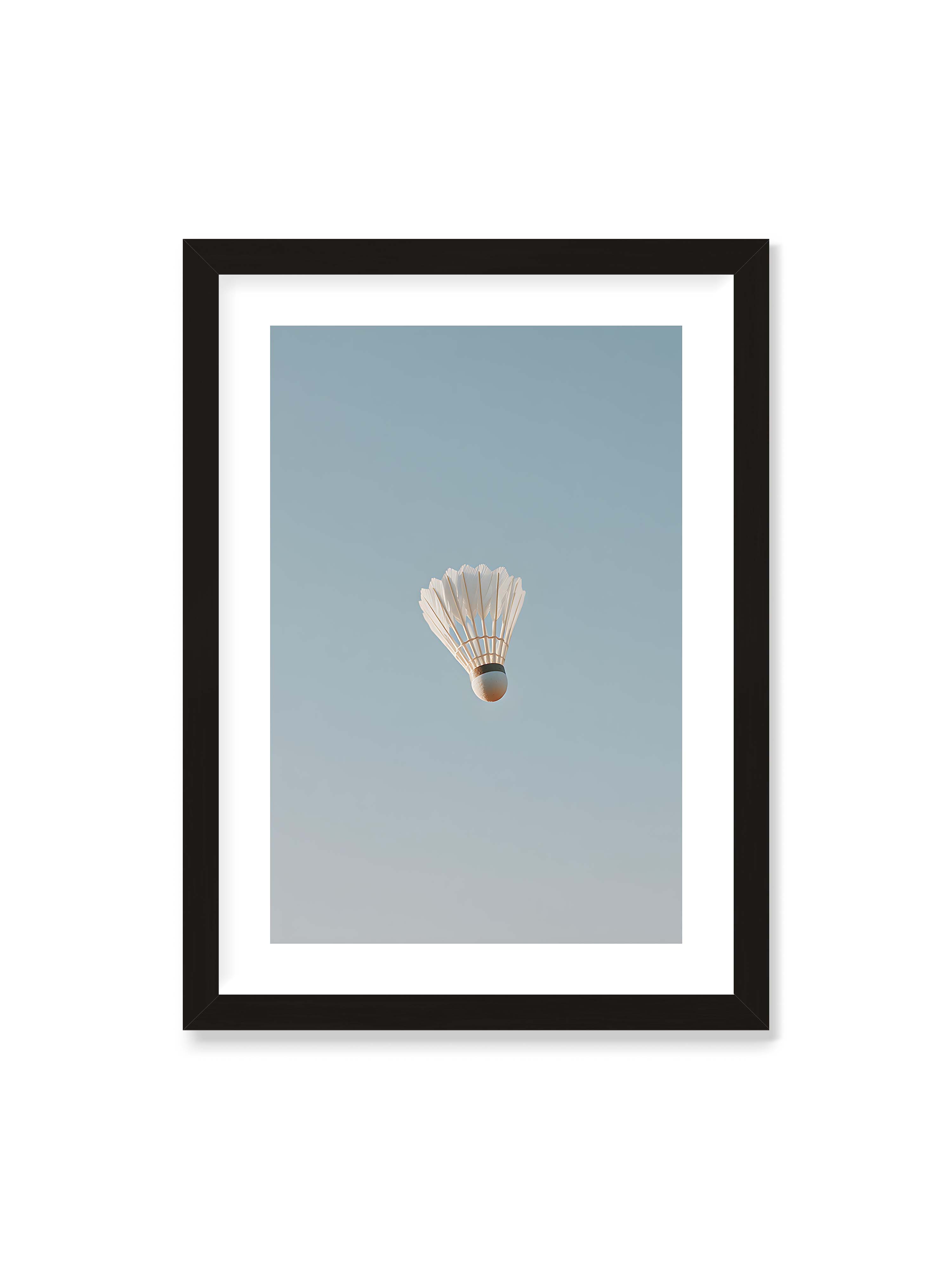 50x70 cm mit weißem Galerie-Rand mit schwarzem Rahmen – Weißer Federball schwebt zentriert vor hellblauem Himmel, minimalistische Komposition mit viel Freiraum.