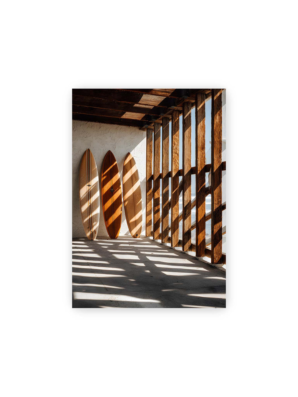 21x30 cm – Drei Surfboards an einer hellen Wand, Holzpfosten werfen diagonale Schatten; Meer und Himmel schimmern im Hintergrund.