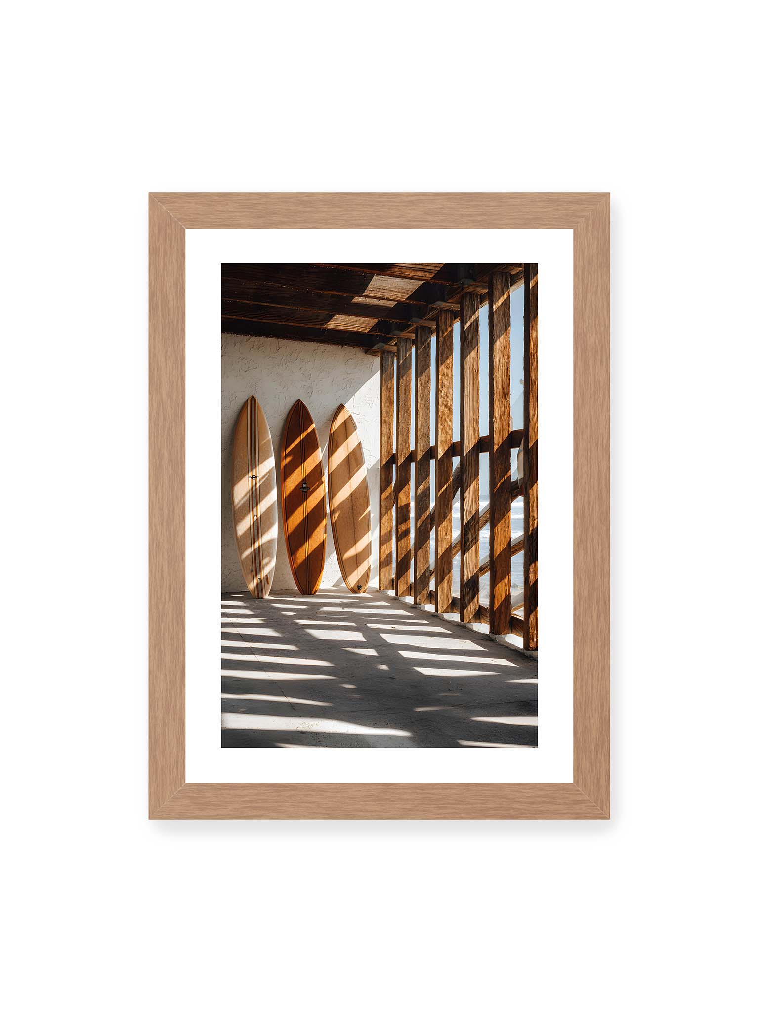 21x30 cm, weißer Galerie-Rand, Eichenrahmen – Drei Surfboards an einer hellen Wand, Holzpfosten werfen diagonale Schatten; Meer und Himmel schimmern im Hintergrund.