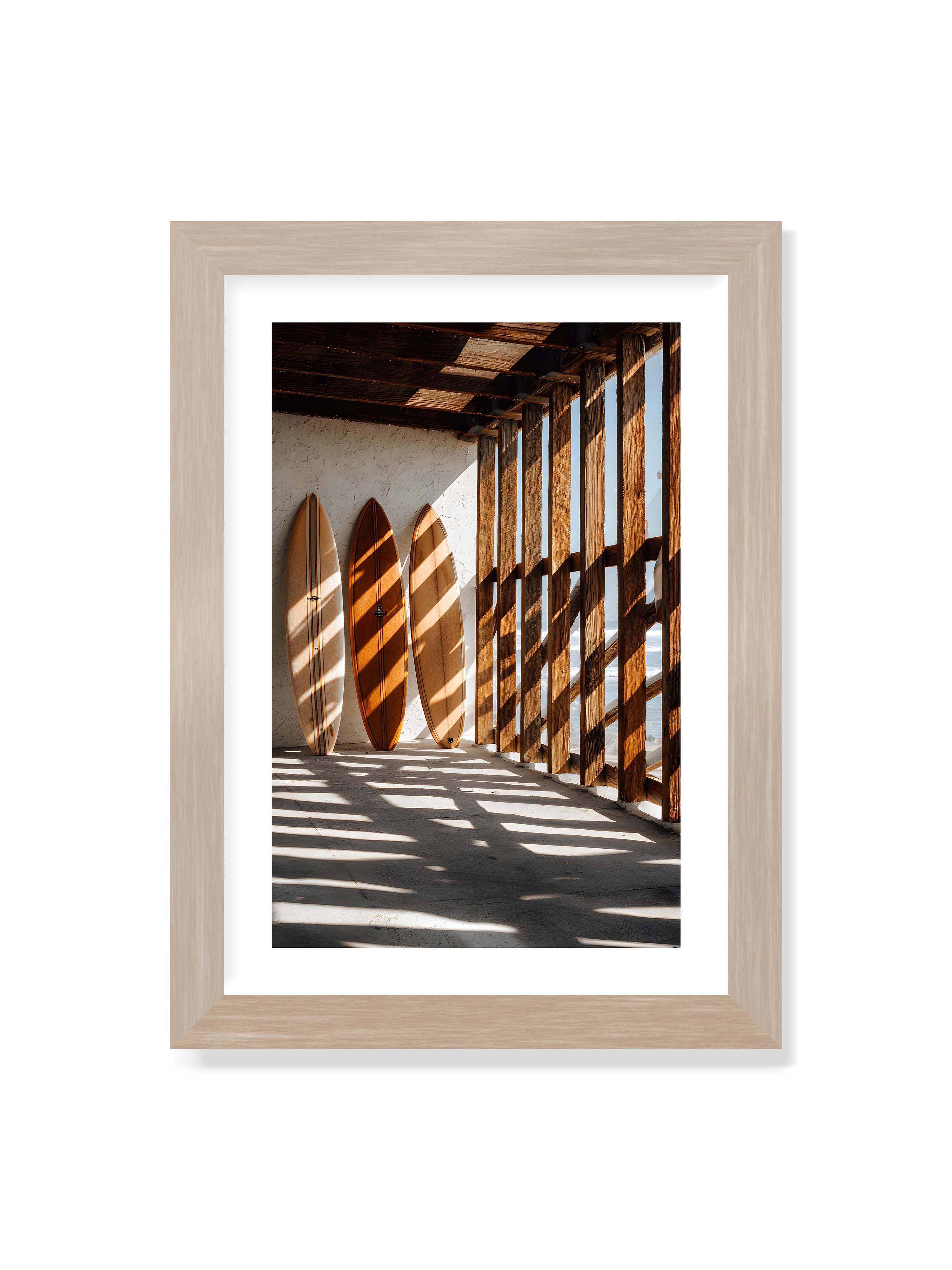 21x30 cm mit weißem Galerie-Rand im Eichenrahmen – Drei Surfboards an einer hellen Wand, Holzpfosten werfen diagonale Schatten; Meer und Himmel schimmern im Hintergrund.