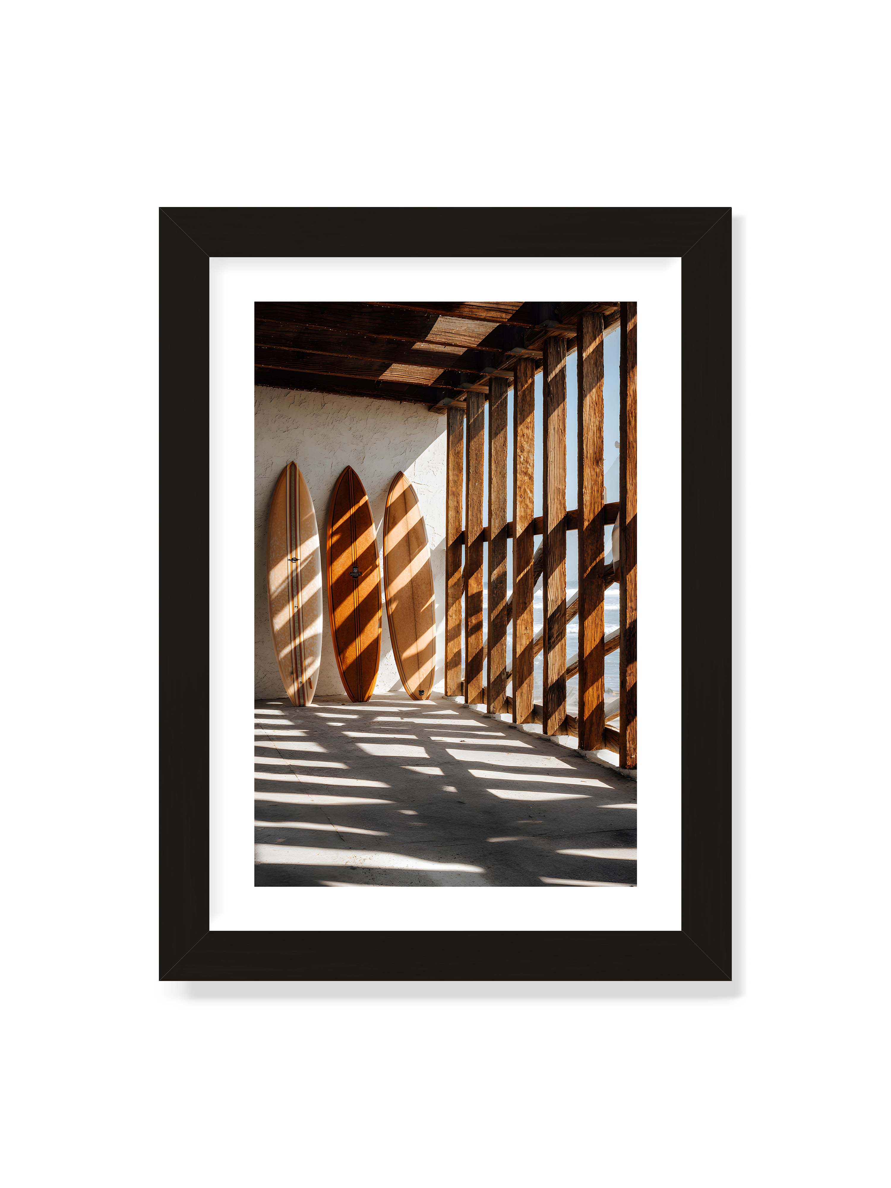 21x30 cm mit weißem Galerie-Rand mit schwarzem Rahmen – Drei Surfboards an einer hellen Wand, Holzpfosten werfen diagonale Schatten; Meer und Himmel schimmern im Hintergrund.