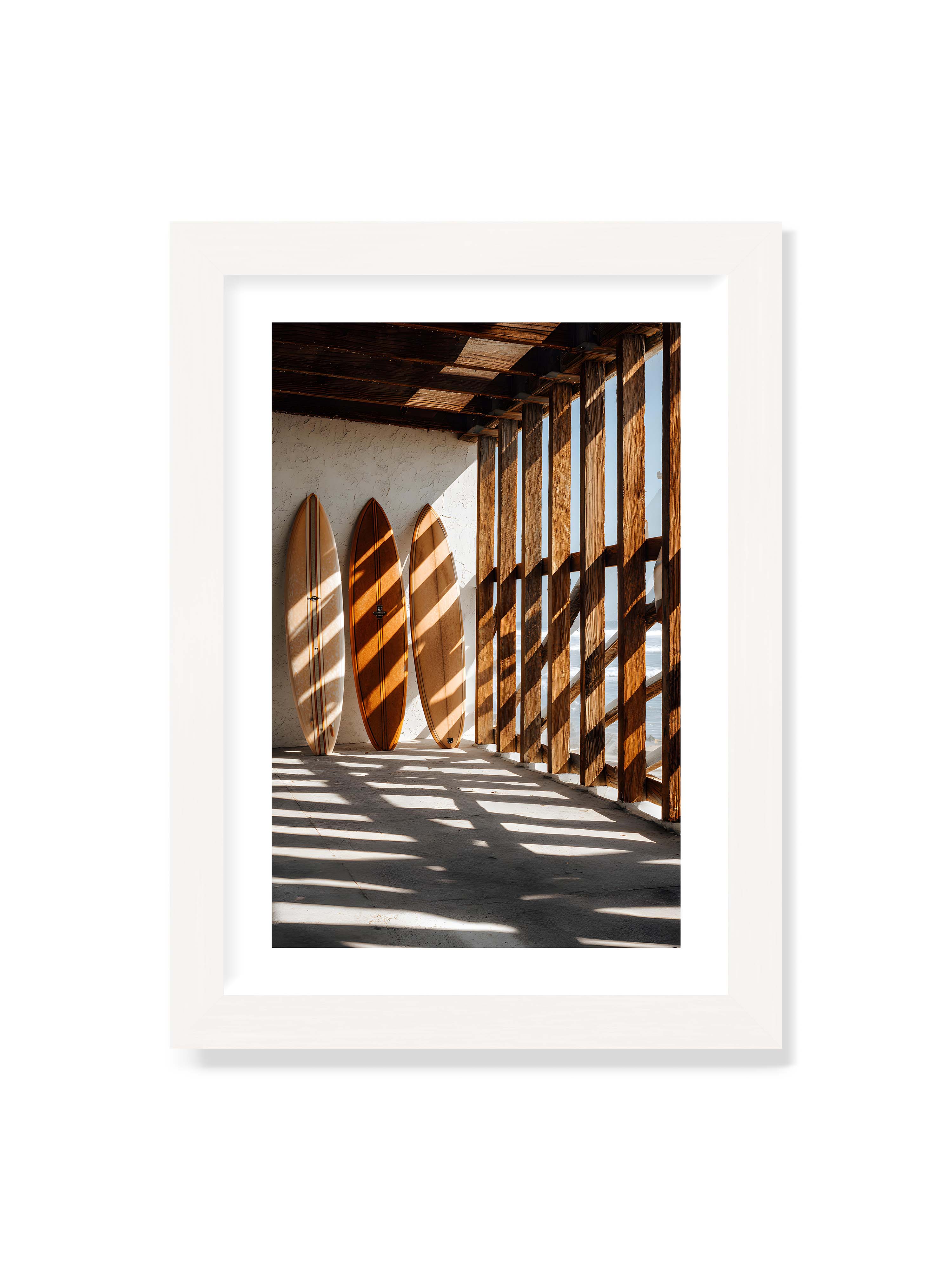 21x30 cm mit weißem Galerie-Rand mit weißem Rahmen – Drei Surfboards an einer hellen Wand, Holzpfosten werfen diagonale Schatten; Meer und Himmel schimmern im Hintergrund.