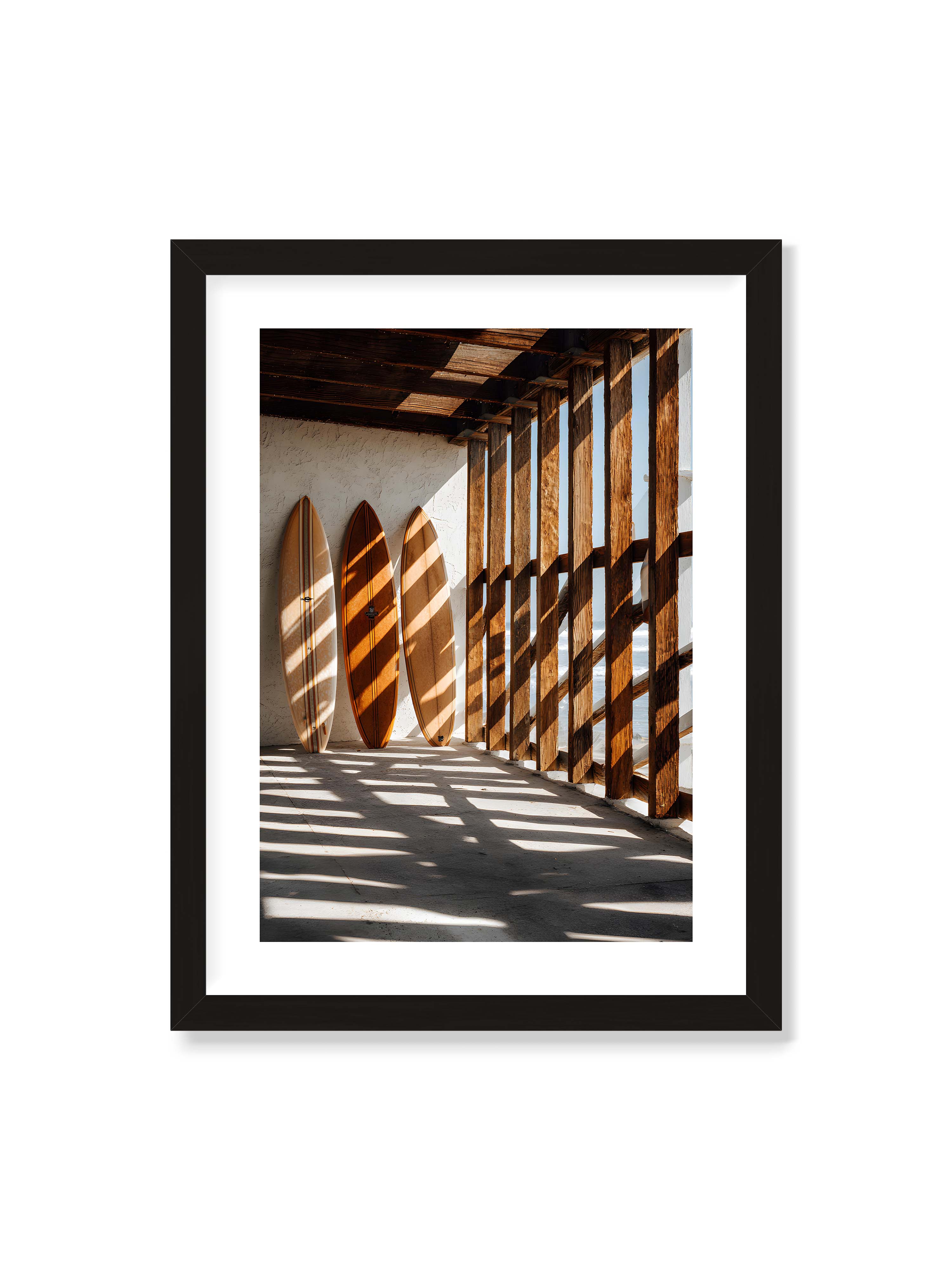 30x40 cm mit weißem Galerie-Rand mit schwarzem Rahmen – Drei Surfboards an einer hellen Wand, Holzpfosten werfen diagonale Schatten; Meer und Himmel schimmern im Hintergrund.