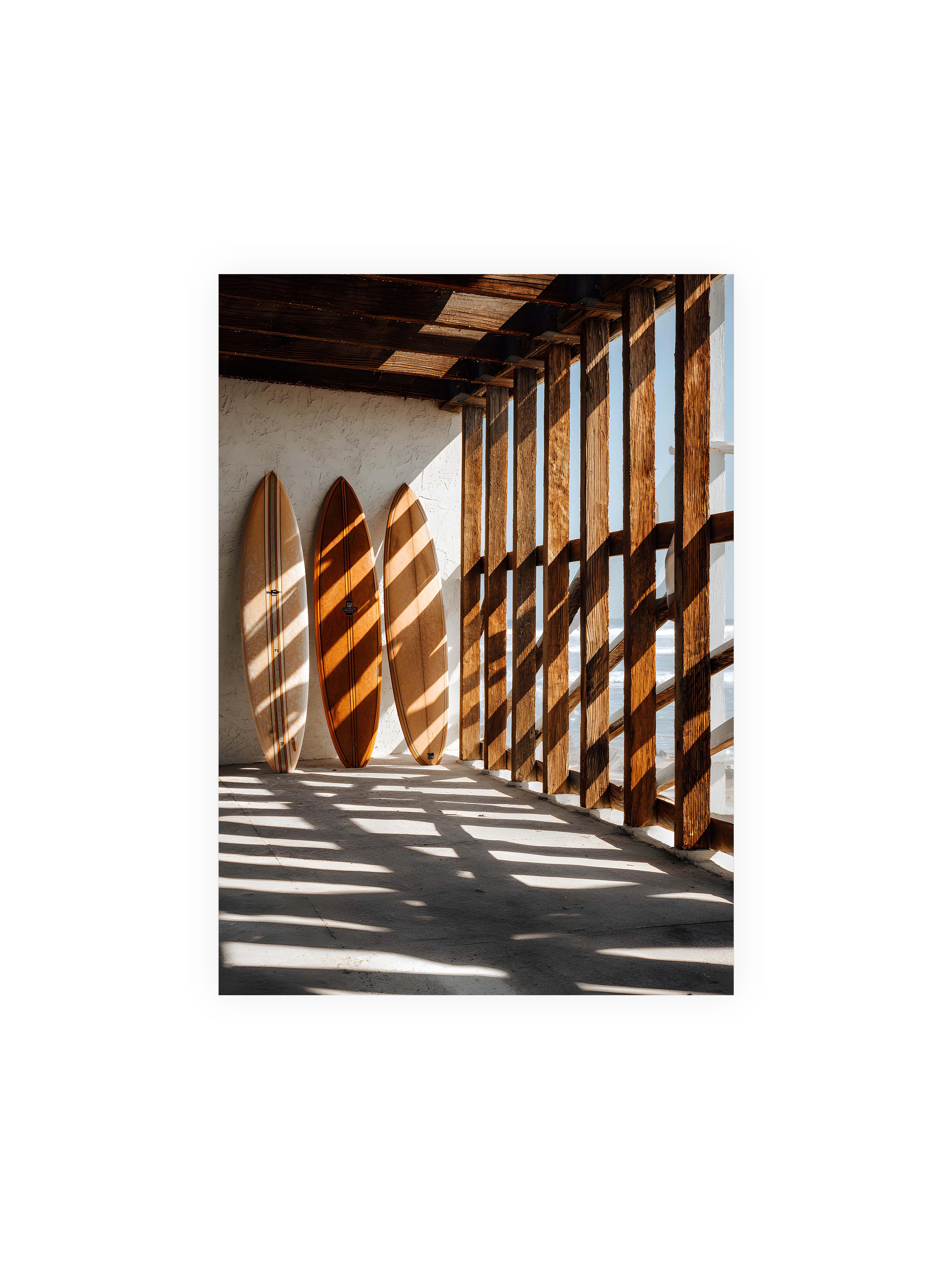 50x70 cm ohne Galerie-Rand ohne Rahmen – Drei Surfboards an einer hellen Wand, Holzpfosten werfen diagonale Schatten; Meer und Himmel schimmern im Hintergrund.