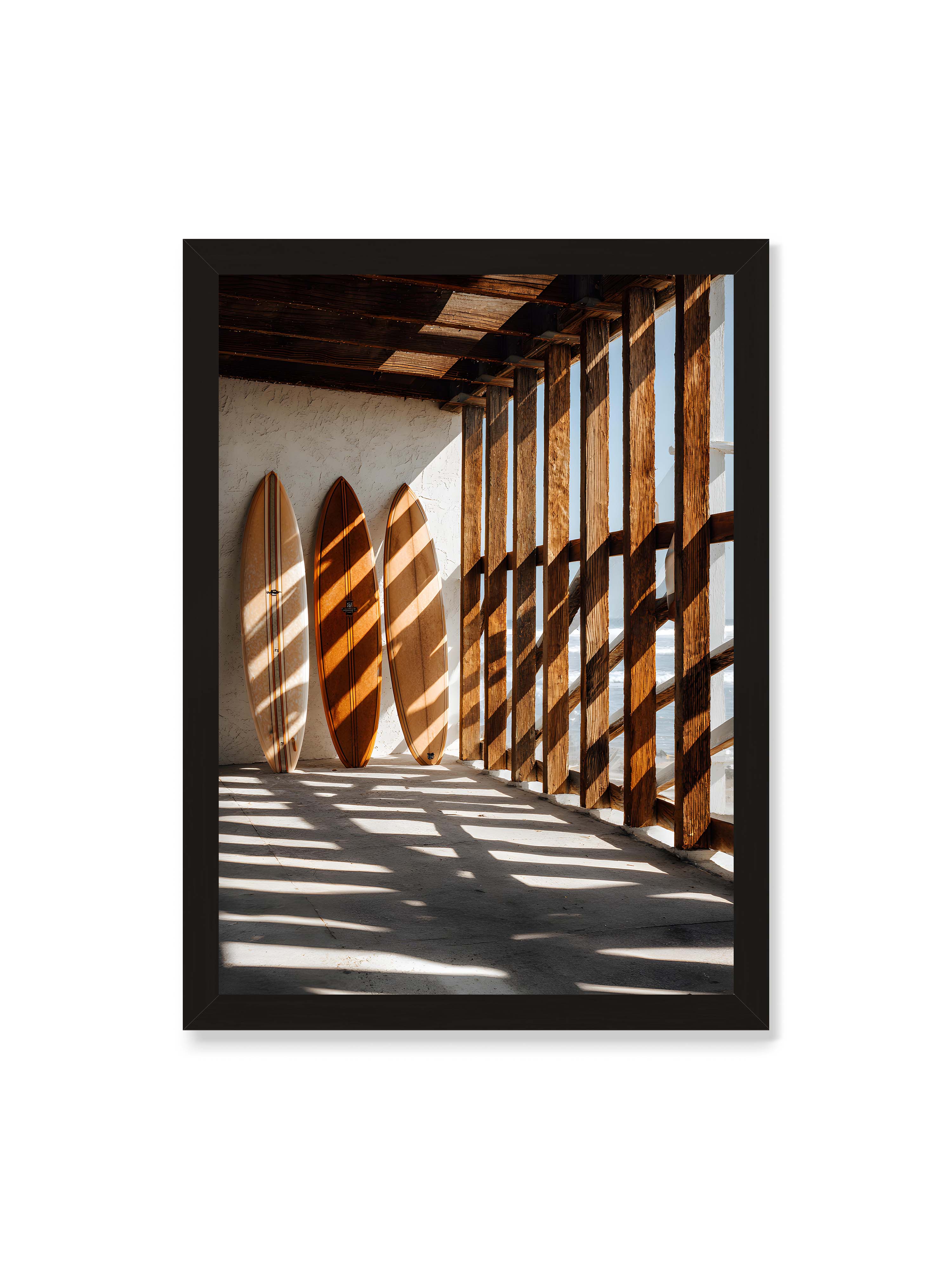 50x70 cm ohne Galerie-Rand mit schwarzem Rahmen – Drei Surfboards an einer hellen Wand, Holzpfosten werfen diagonale Schatten; Meer und Himmel schimmern im Hintergrund.