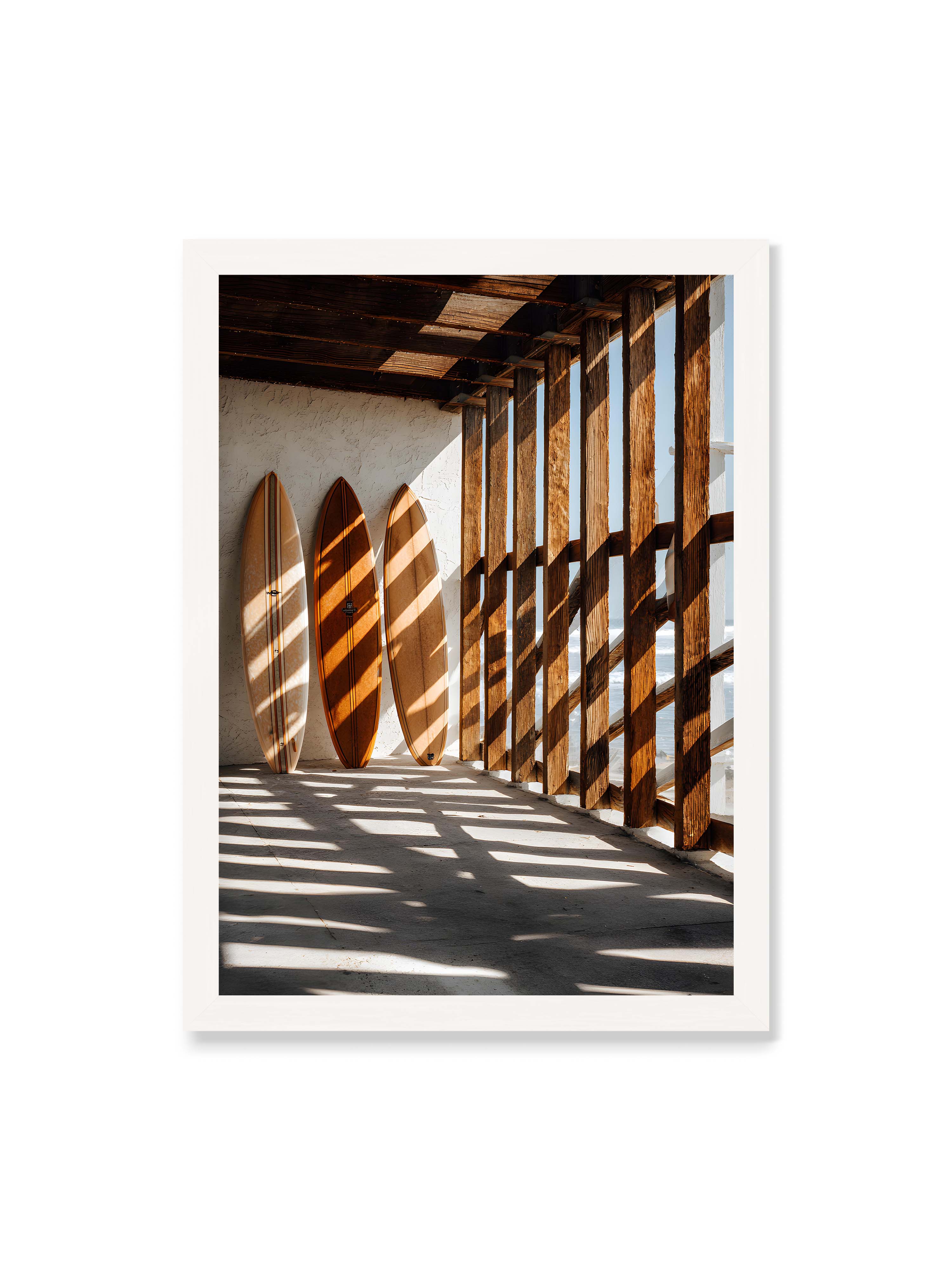 50x70 cm ohne Galerie-Rand mit weißem Rahmen – Drei Surfboards an einer hellen Wand, Holzpfosten werfen diagonale Schatten; Meer und Himmel schimmern im Hintergrund.