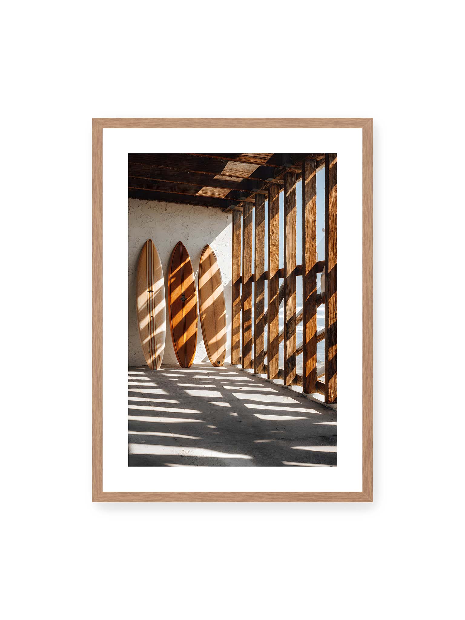 50x70 cm, weißer Galerie-Rand, Eichenrahmen – Drei Surfboards an einer hellen Wand, Holzpfosten werfen diagonale Schatten; Meer und Himmel schimmern im Hintergrund.