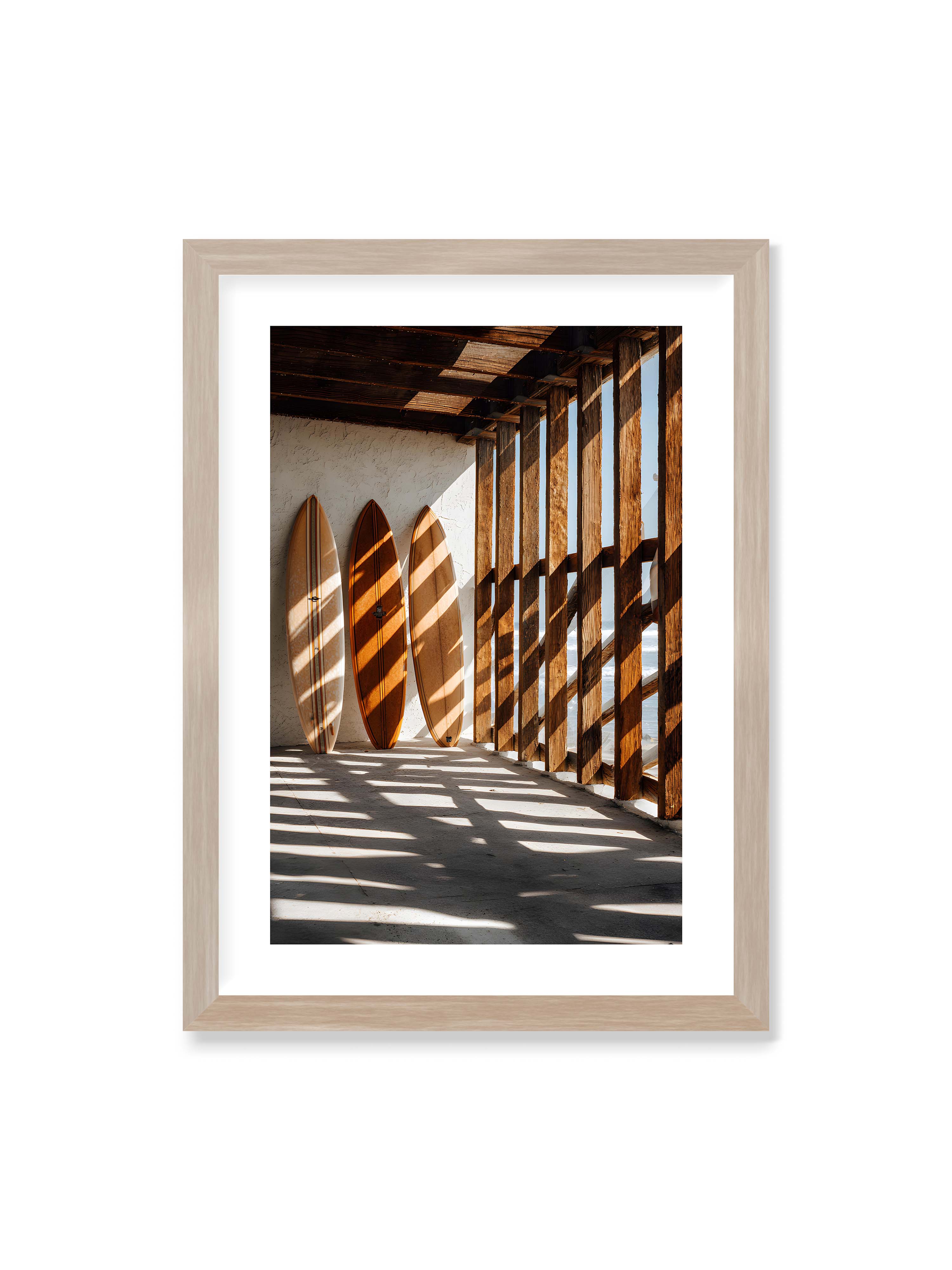 50x70 cm mit weißem Galerie-Rand im Eichenrahmen – Drei Surfboards an einer hellen Wand, Holzpfosten werfen diagonale Schatten; Meer und Himmel schimmern im Hintergrund.