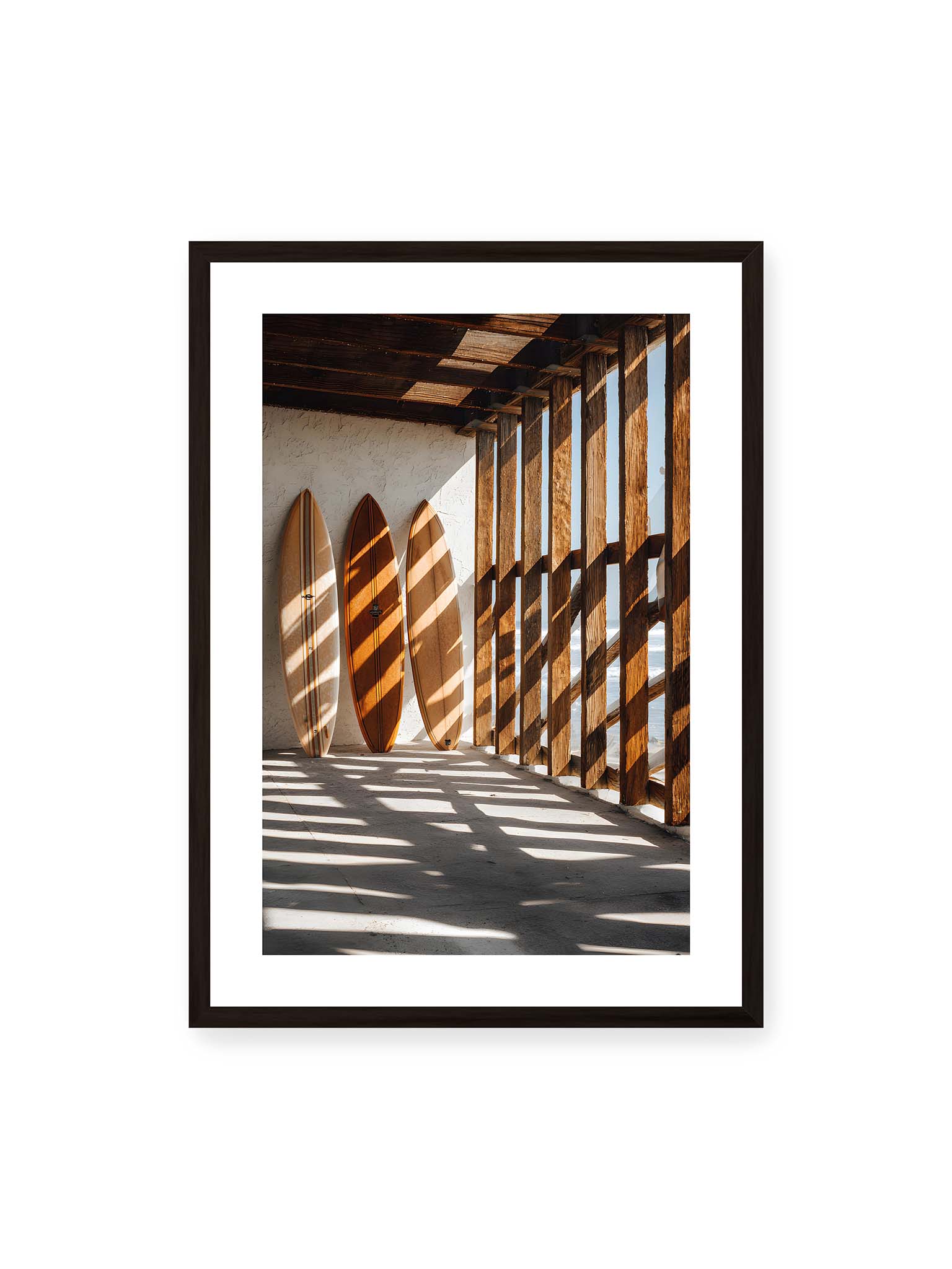50x70 cm, weißer Galerie-Rand, schwarzer Rahmen – Drei Surfboards an einer hellen Wand, Holzpfosten werfen diagonale Schatten; Meer und Himmel schimmern im Hintergrund.