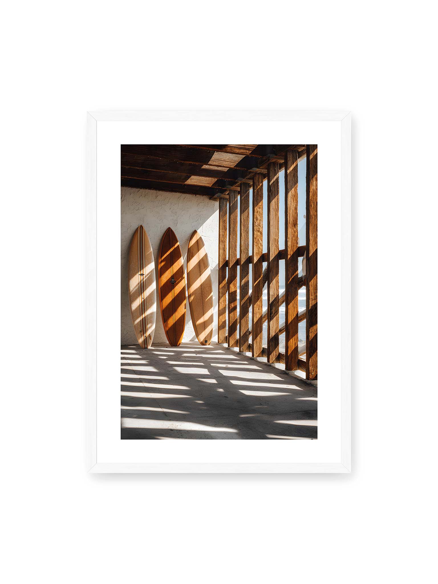 50x70 cm, weißer Galerie-Rand, weißer Rahmen – Drei Surfboards an einer hellen Wand, Holzpfosten werfen diagonale Schatten; Meer und Himmel schimmern im Hintergrund.