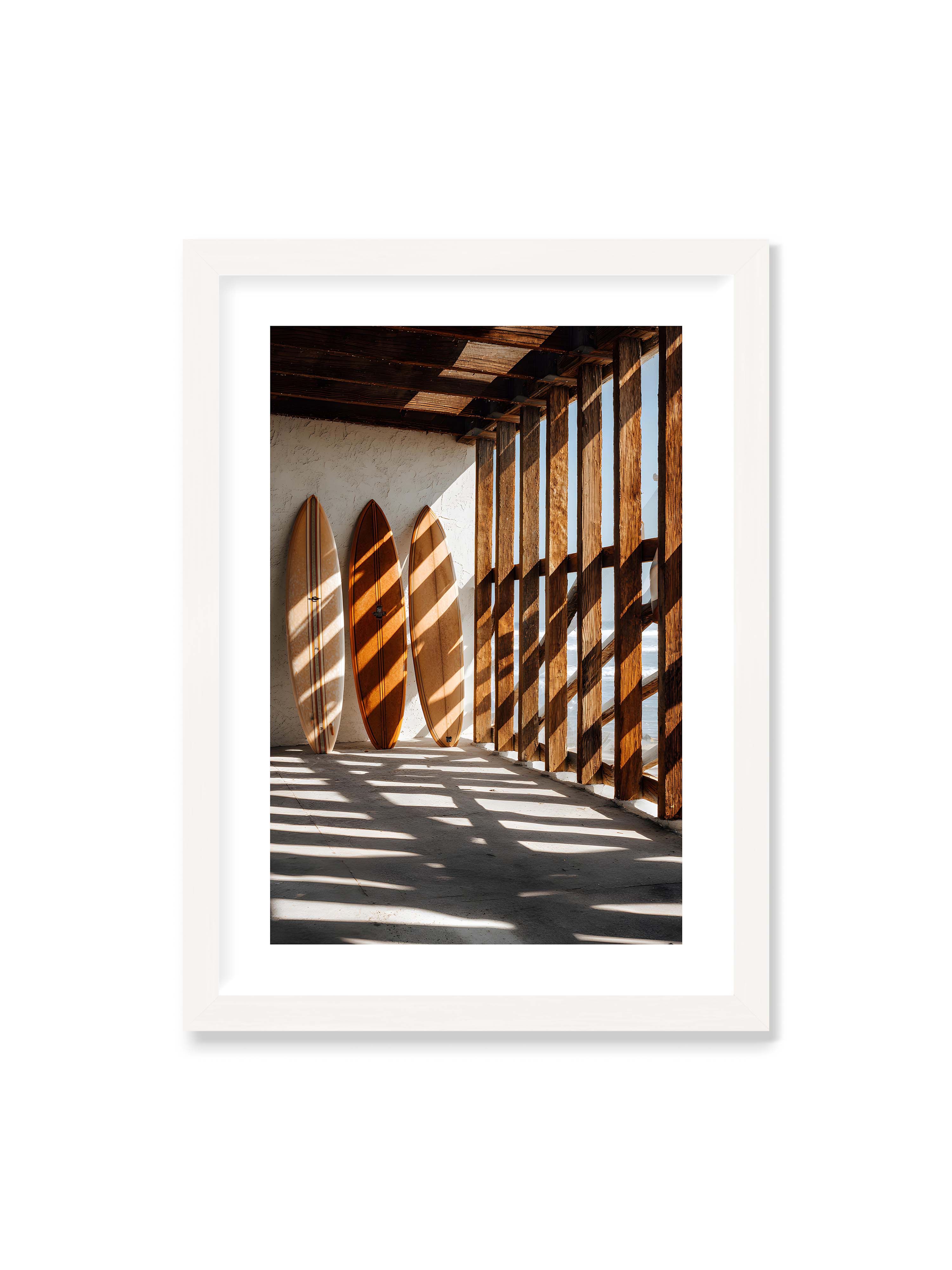 50x70 cm mit weißem Galerie-Rand mit weißem Rahmen – Drei Surfboards an einer hellen Wand, Holzpfosten werfen diagonale Schatten; Meer und Himmel schimmern im Hintergrund.