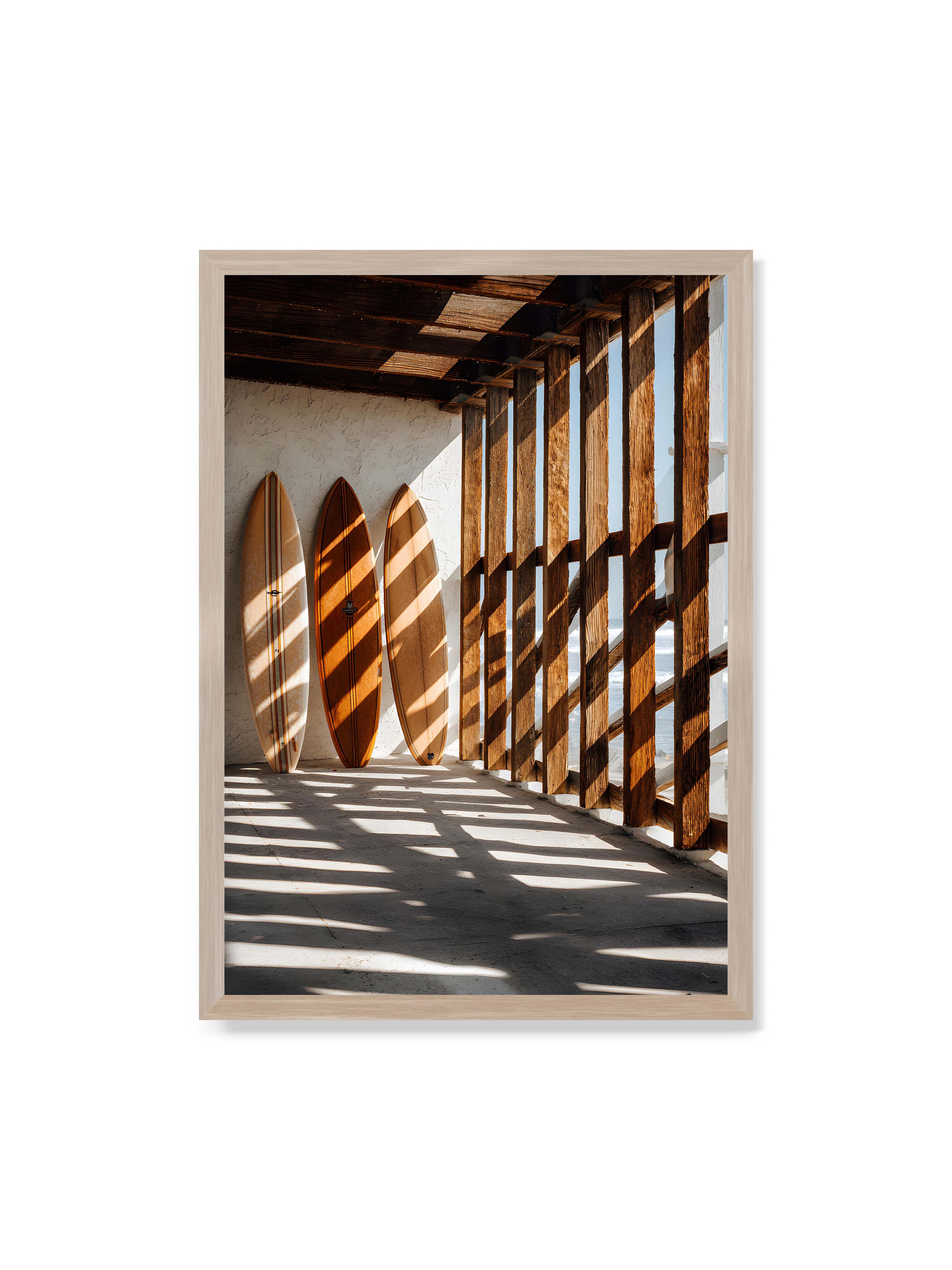 70x100 cm ohne Galerie-Rand im Eichenrahmen – Drei Surfboards an einer hellen Wand, Holzpfosten werfen diagonale Schatten; Meer und Himmel schimmern im Hintergrund.