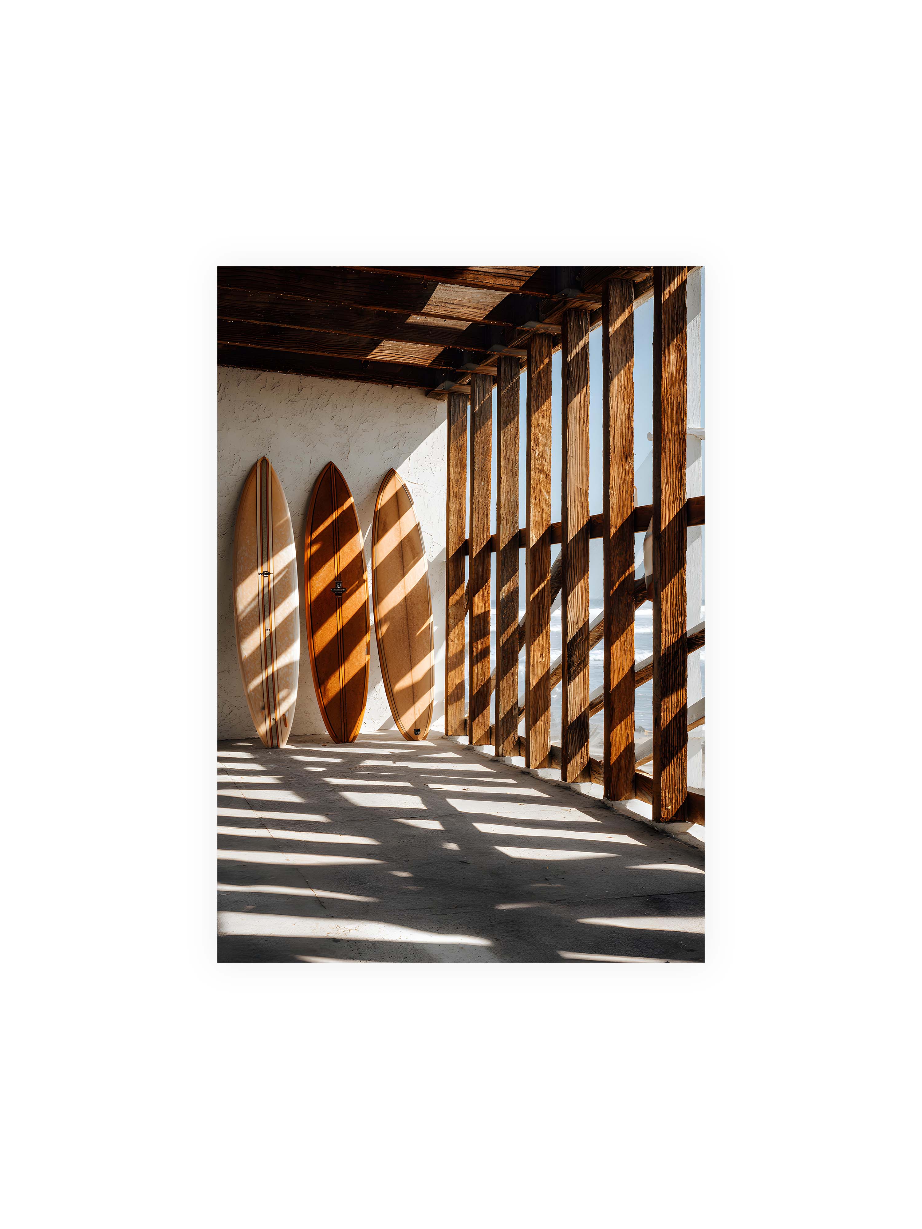 70x100 cm ohne Galerie-Rand ohne Rahmen – Drei Surfboards an einer hellen Wand, Holzpfosten werfen diagonale Schatten; Meer und Himmel schimmern im Hintergrund.