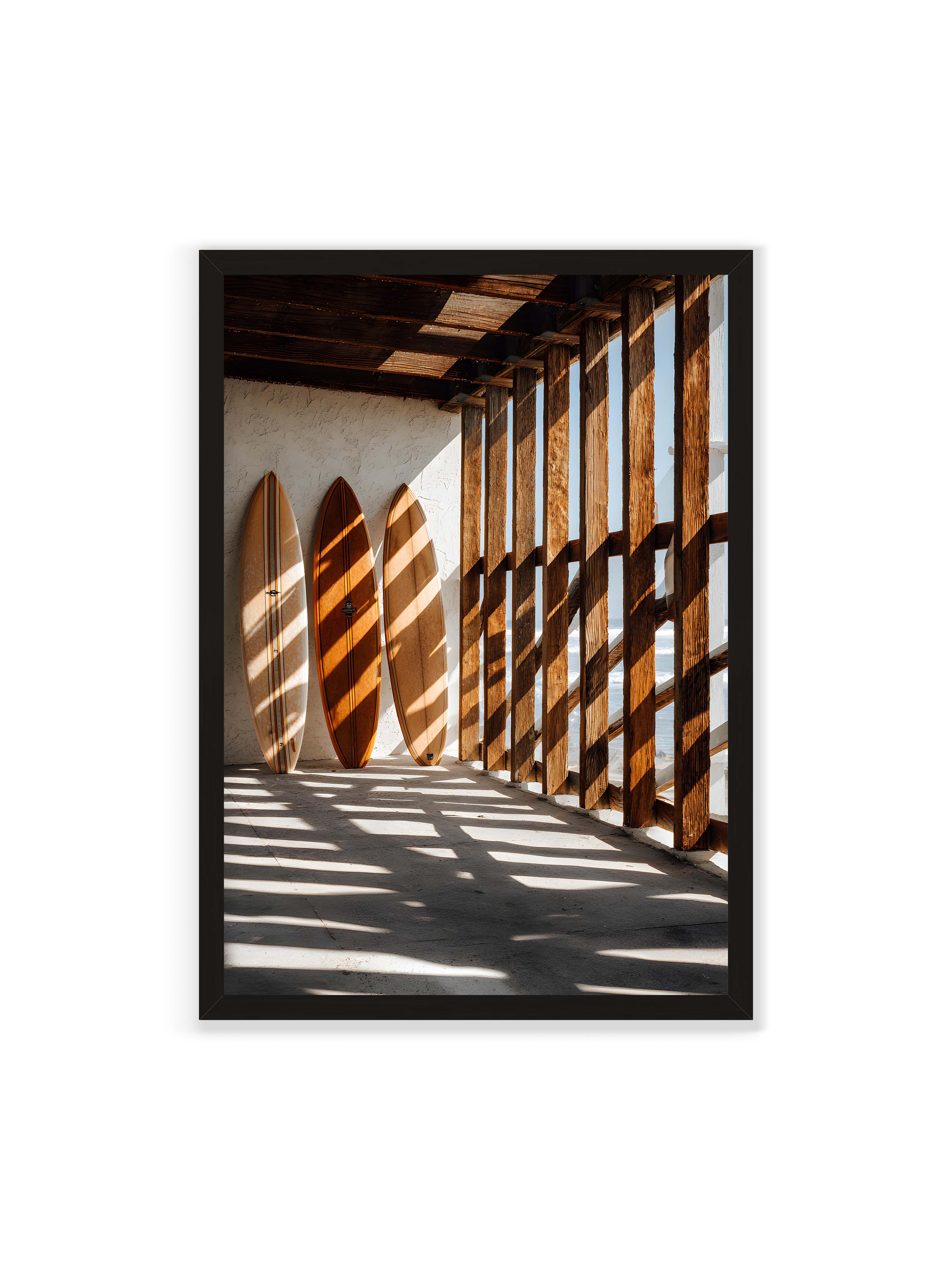 70x100 cm ohne Galerie-Rand mit schwarzem Rahmen – Drei Surfboards an einer hellen Wand, Holzpfosten werfen diagonale Schatten; Meer und Himmel schimmern im Hintergrund.