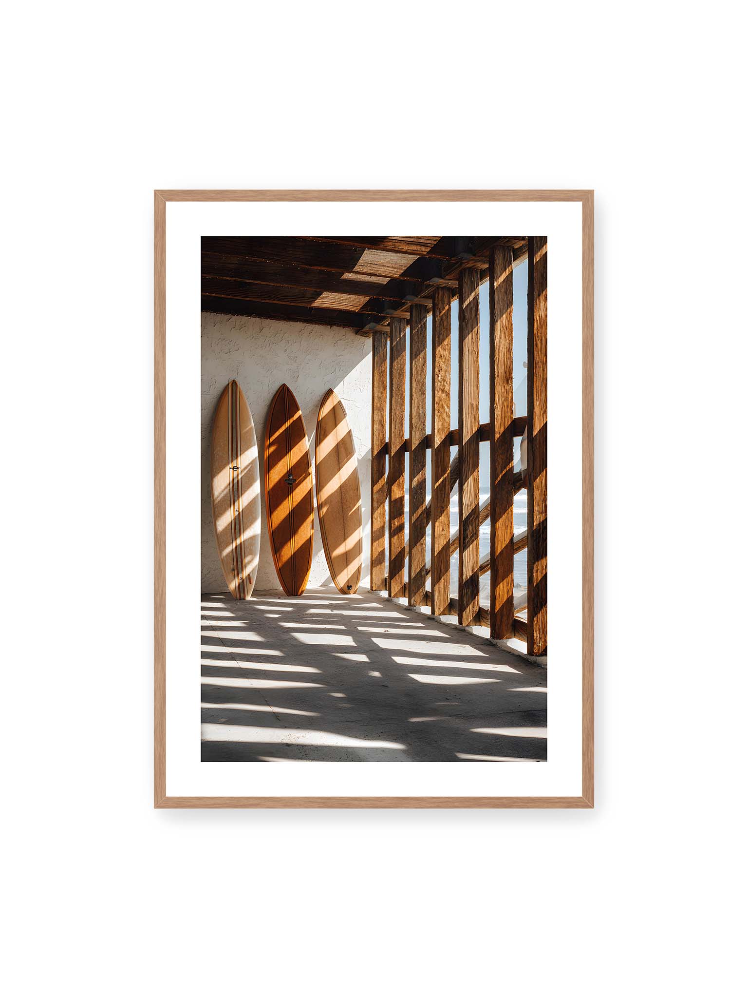 70x100 cm, weißer Galerie-Rand, Eichenrahmen – Drei Surfboards an einer hellen Wand, Holzpfosten werfen diagonale Schatten; Meer und Himmel schimmern im Hintergrund.