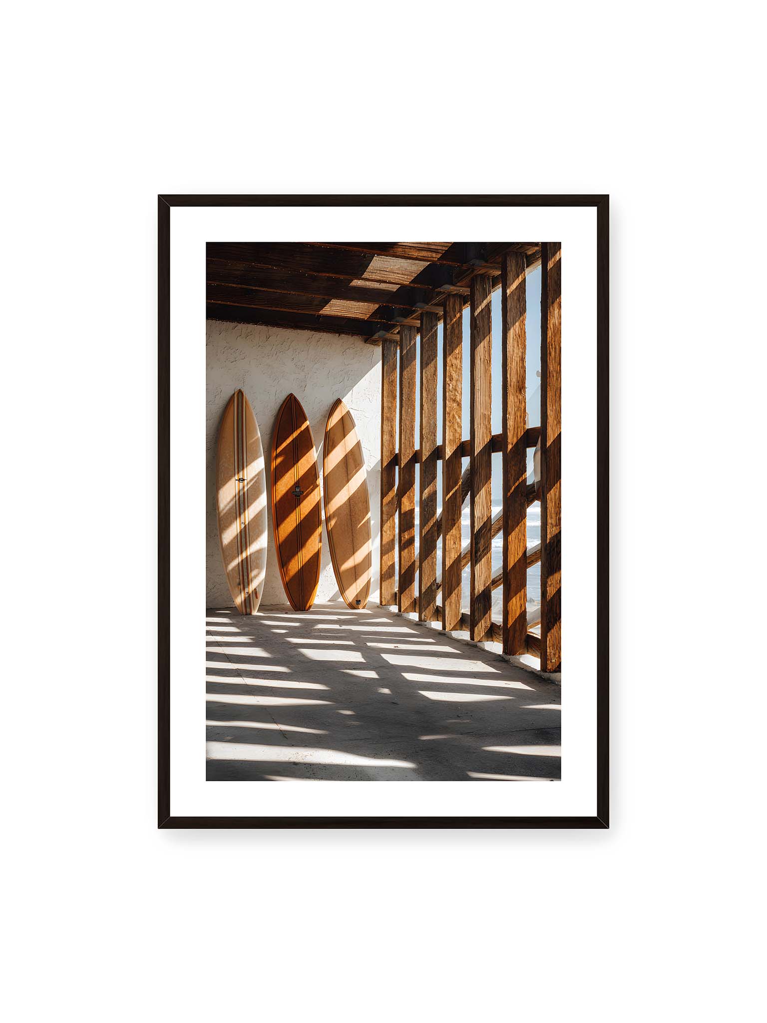70x100 cm, weißer Galerie-Rand, schwarzer Rahmen – Drei Surfboards an einer hellen Wand, Holzpfosten werfen diagonale Schatten; Meer und Himmel schimmern im Hintergrund.
