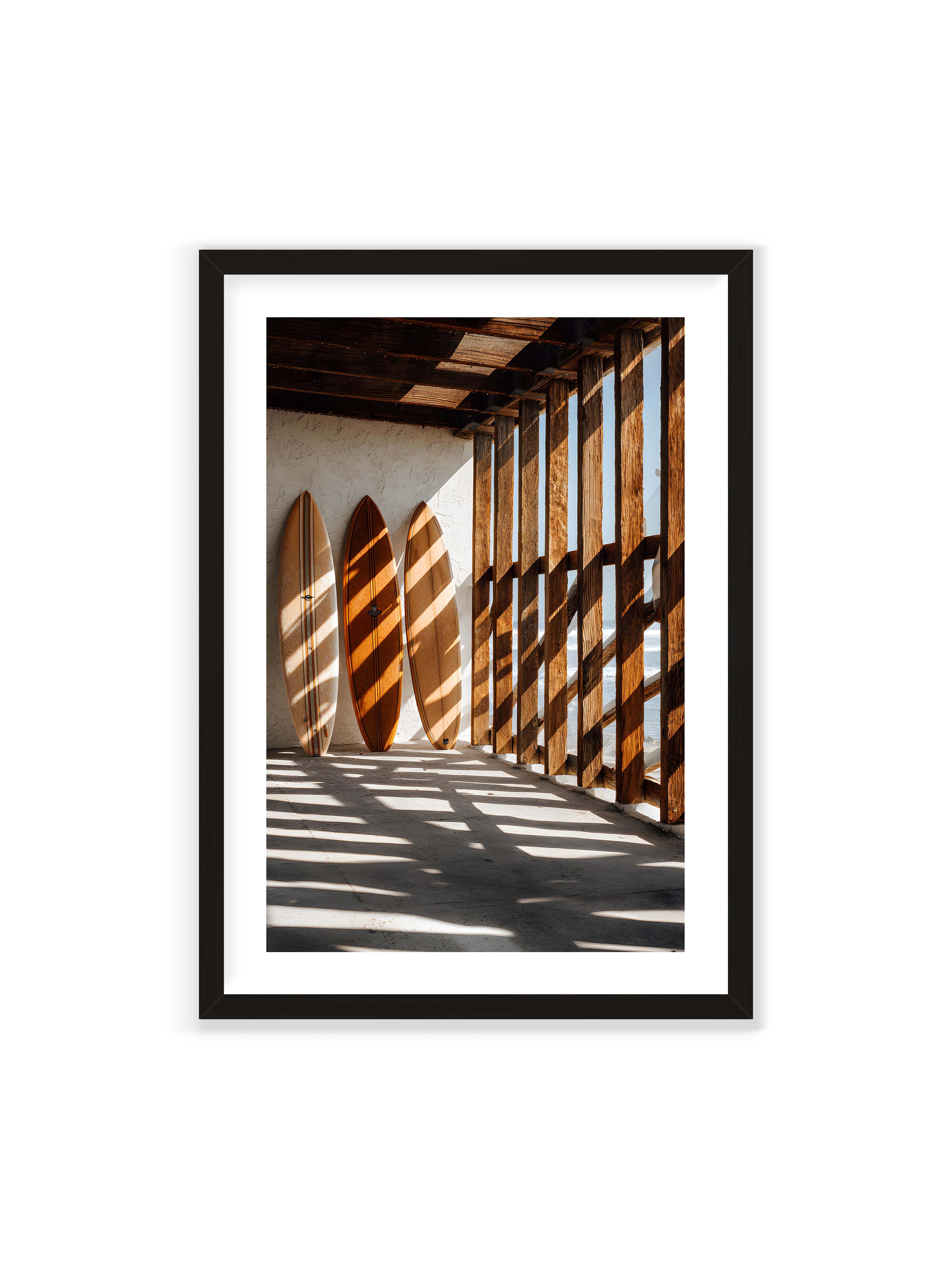 70x100 cm mit weißem Galerie-Rand mit schwarzem Rahmen – Drei Surfboards an einer hellen Wand, Holzpfosten werfen diagonale Schatten; Meer und Himmel schimmern im Hintergrund.