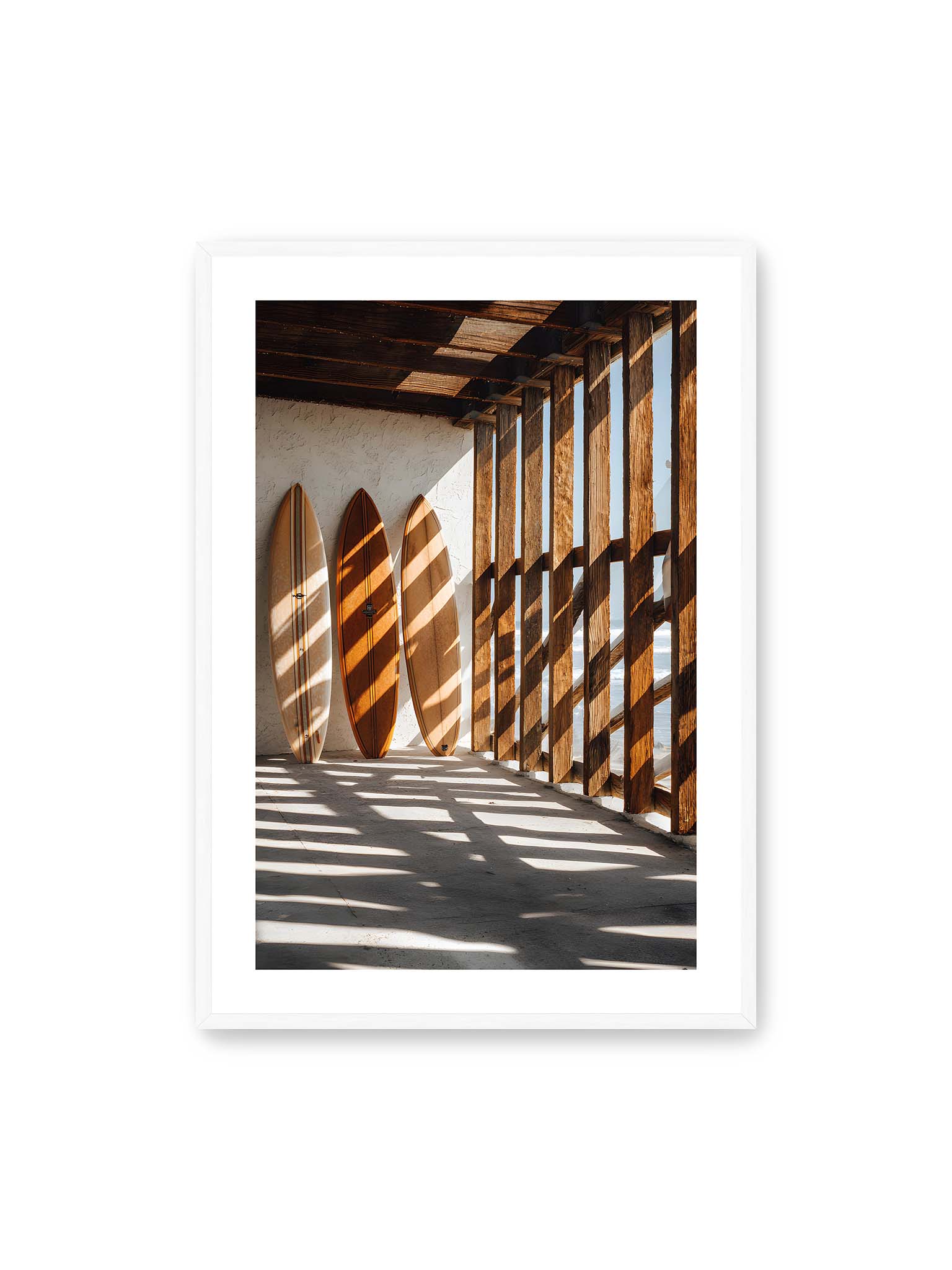 70x100 cm, weißer Galerie-Rand, weißer Rahmen – Drei Surfboards an einer hellen Wand, Holzpfosten werfen diagonale Schatten; Meer und Himmel schimmern im Hintergrund.