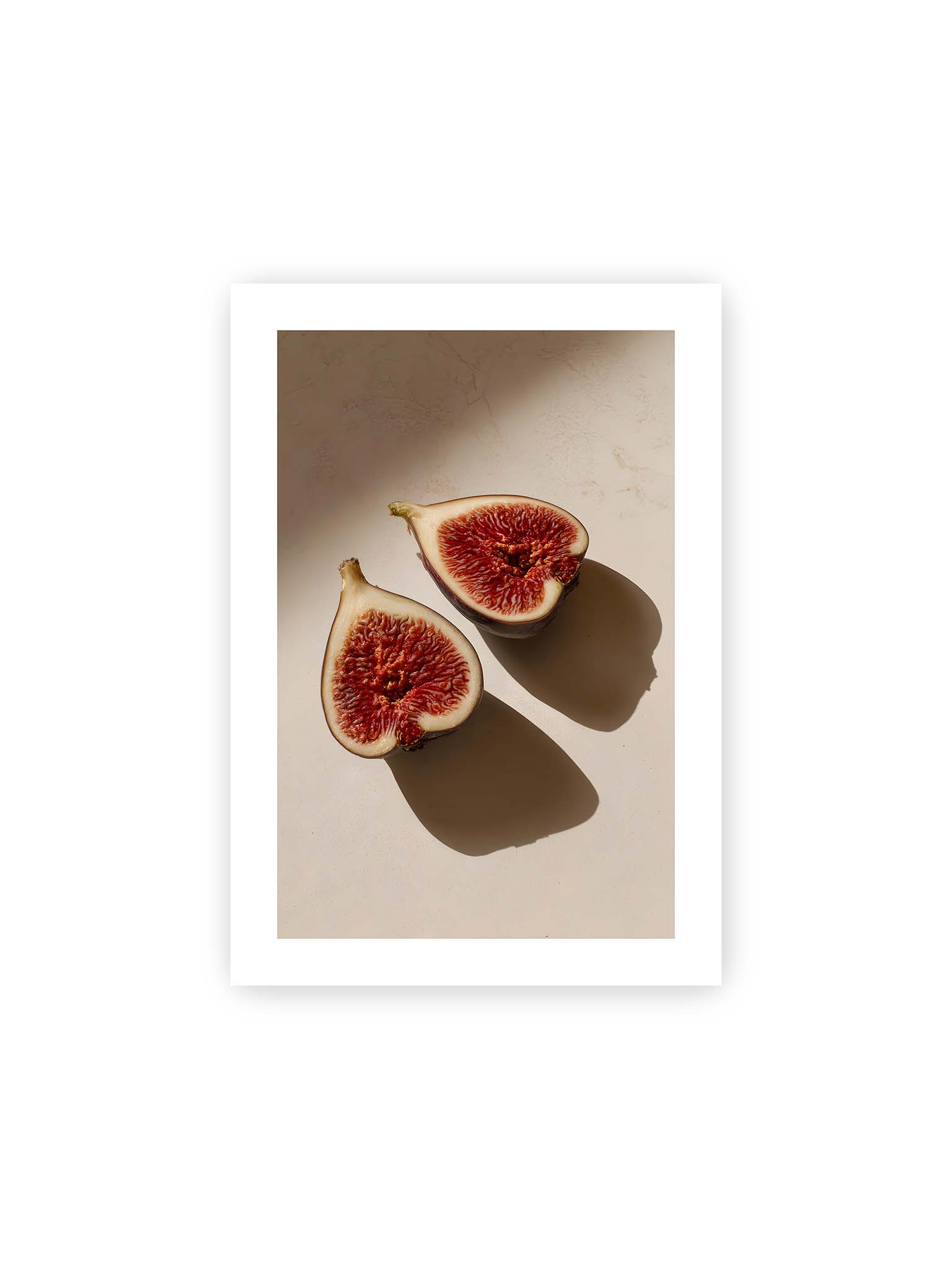 21x30 cm, weißer Galerie-Rand – Halbierte Feigen auf heller Oberfläche mit warmem Licht und langen Schatten, sichtbar strukturierte rote Fruchtmitte.