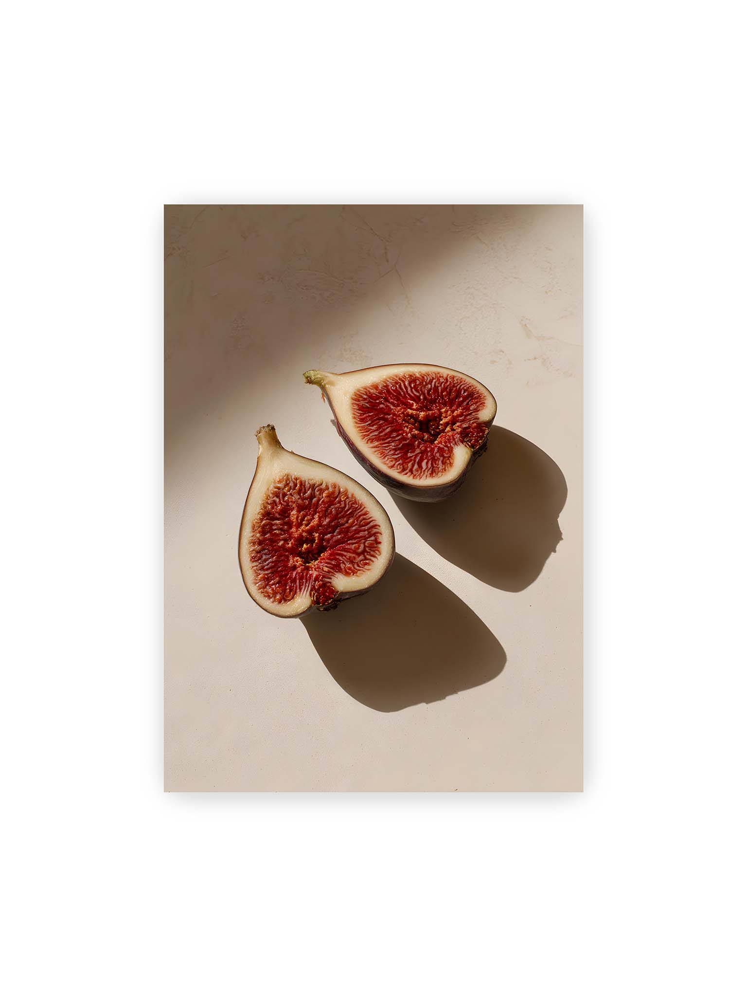 50x70 cm – Halbierte Feigen auf heller Oberfläche mit warmem Licht und langen Schatten, sichtbar strukturierte rote Fruchtmitte.