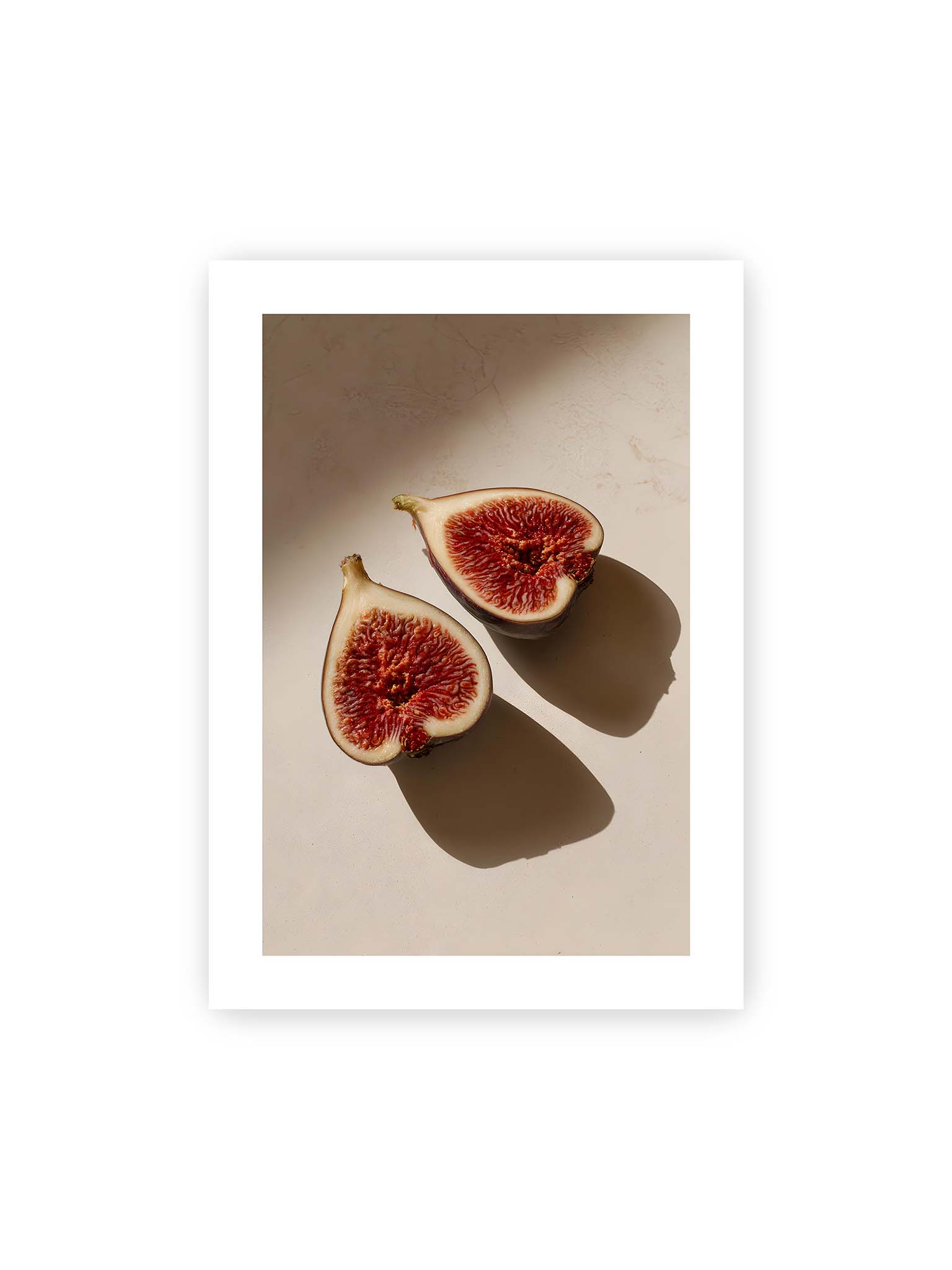 50x70 cm, weißer Galerie-Rand – Halbierte Feigen auf heller Oberfläche mit warmem Licht und langen Schatten, sichtbar strukturierte rote Fruchtmitte.