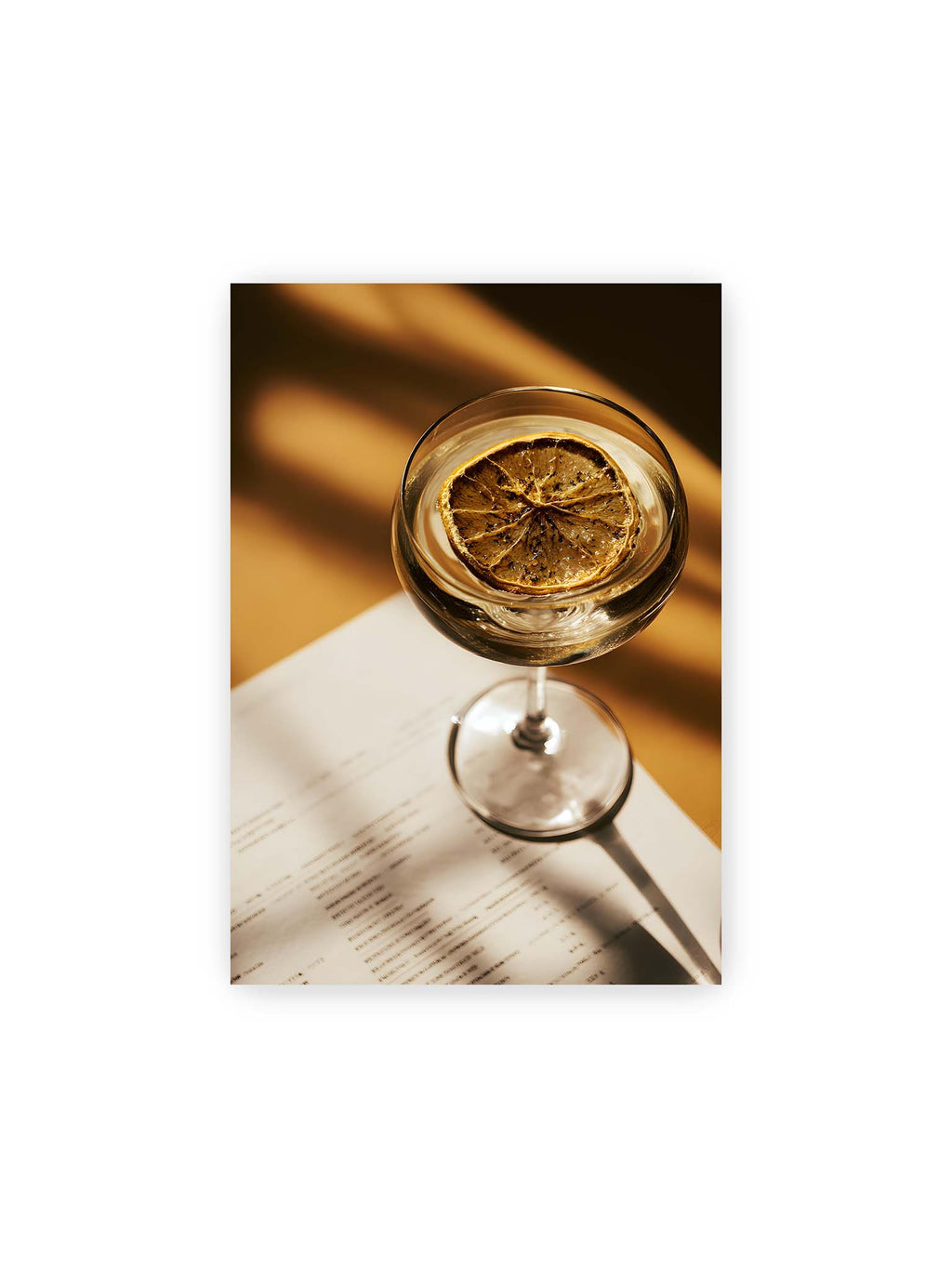 21x30 cm – Cocktailglas mit klarem Getränk und getrockneter Zitrusscheibe von oben fotografiert, warmes Licht und Schatten auf Tisch und Papier.