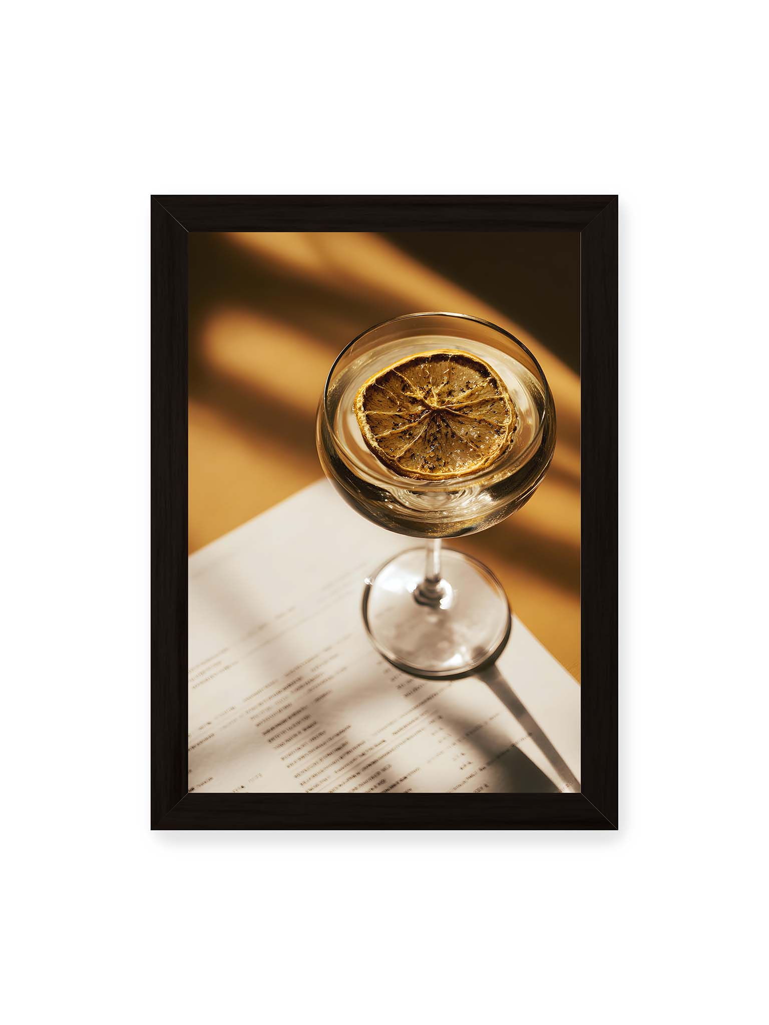 21x30 cm, schwarzer Rahmen – Cocktailglas mit klarem Getränk und getrockneter Zitrusscheibe von oben fotografiert, warmes Licht und Schatten auf Tisch und Papier.