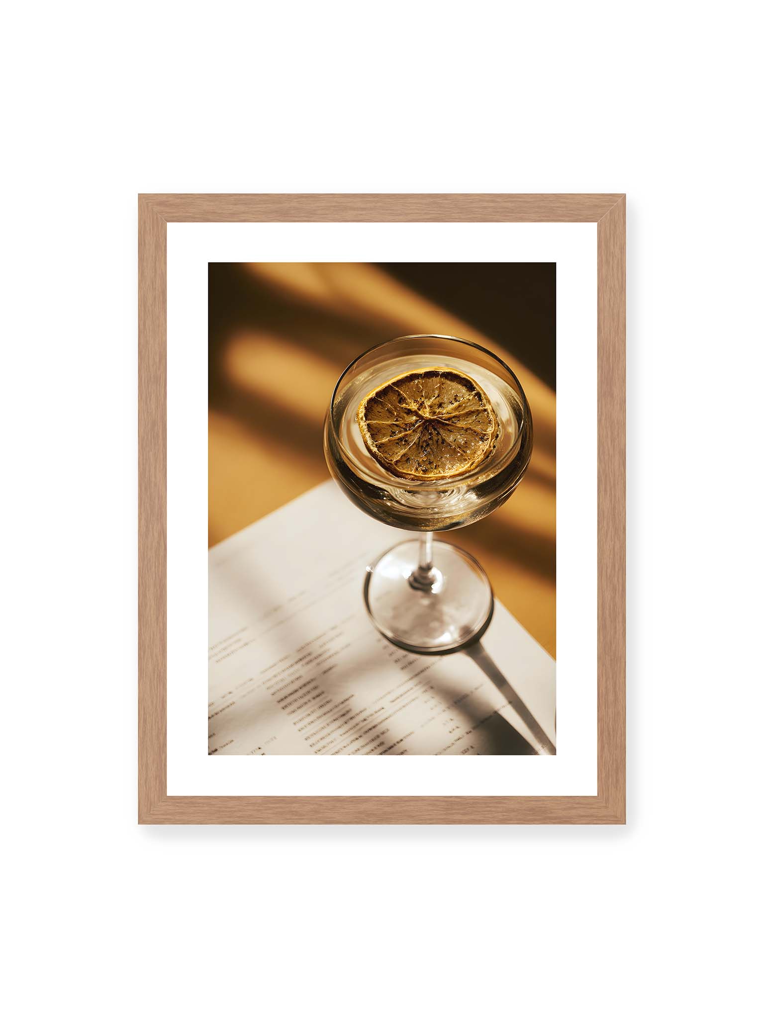 30x40 cm, weißer Galerie-Rand, Eichenrahmen – Cocktailglas mit klarem Getränk und getrockneter Zitrusscheibe von oben fotografiert, warmes Licht und Schatten auf Tisch und Papier.