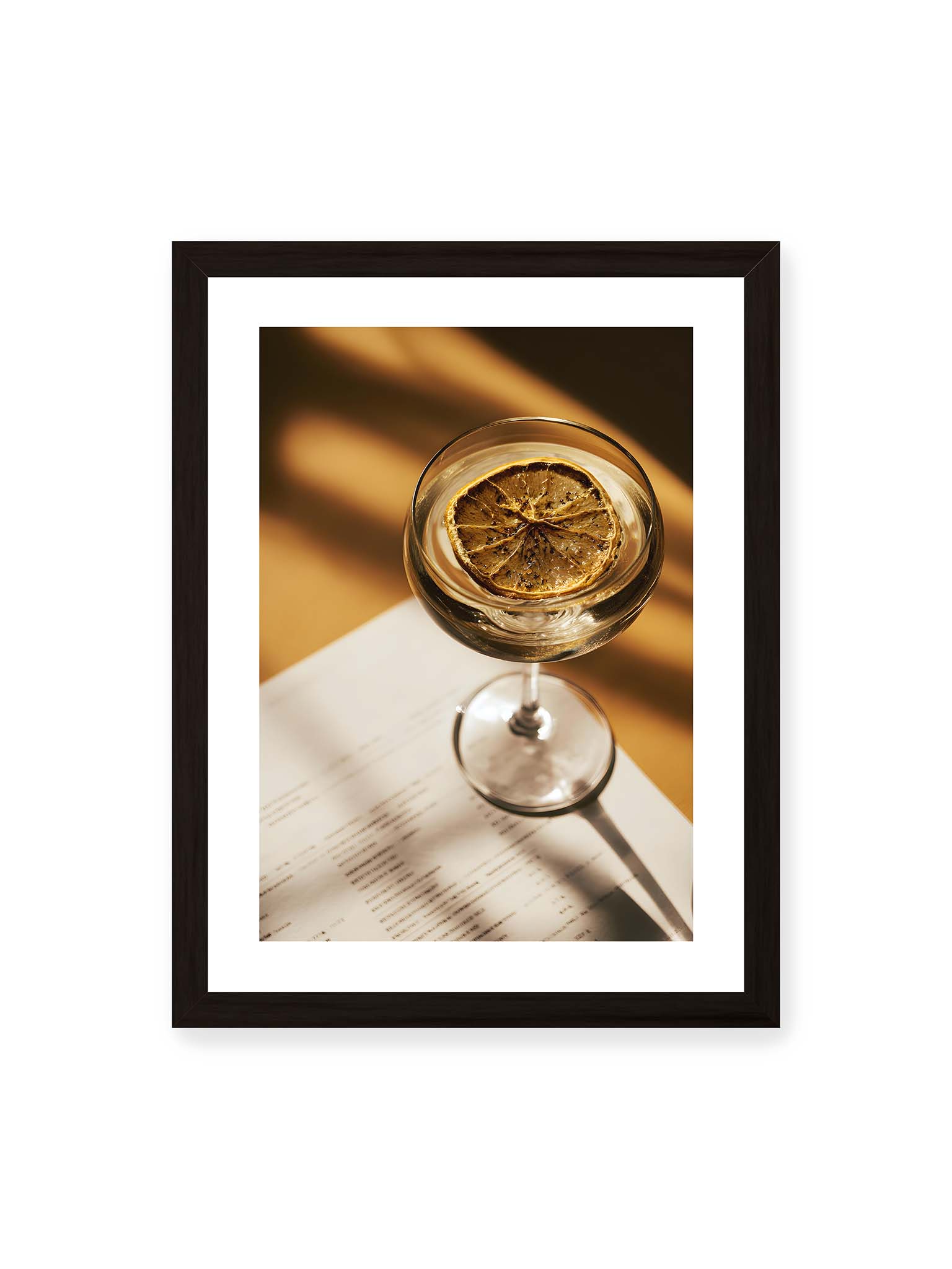 30x40 cm, weißer Galerie-Rand, schwarzer Rahmen – Cocktailglas mit klarem Getränk und getrockneter Zitrusscheibe von oben fotografiert, warmes Licht und Schatten auf Tisch und Papier.